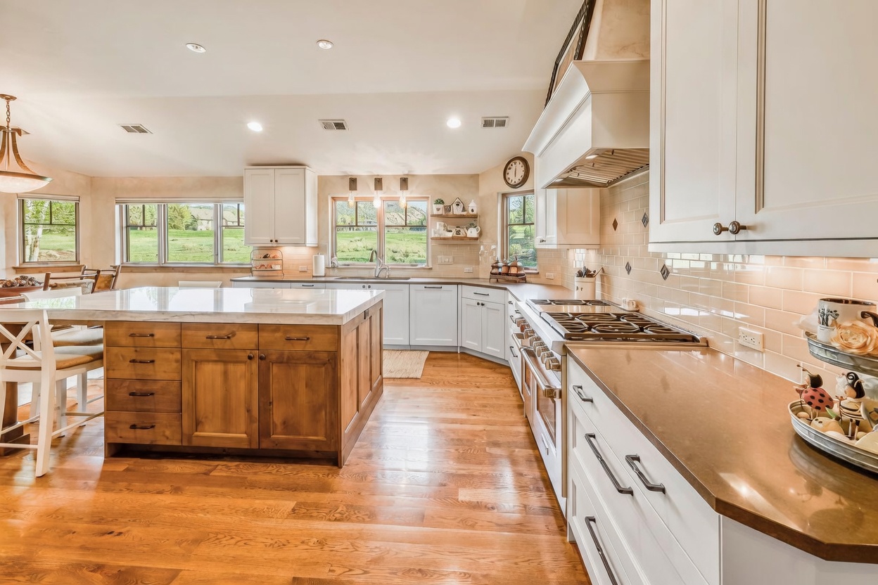 This bright and spacious kitchen features a large central island with a light-toned stone countertop and warm-toned wood cabinetry. The perimeter includes white cabinets, a professional-style gas range with a custom hood, and a light-colored subway tile backsplash. Large windows above the sink provide a scenic view of the surrounding landscape, creating an inviting and airy atmosphere.