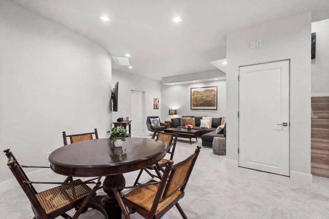 This open-concept living and dining area features a neutral color palette and a blend of traditional and contemporary furnishings. In the foreground, a round dark wood dining table is surrounded by four cane-back chairs, while the background showcases a comfortable seating area with a dark sectional sofa and a framed landscape print. The space is illuminated by recessed lighting and includes a staircase leading upward, creating a functional and inviting multi-purpose living environment.
