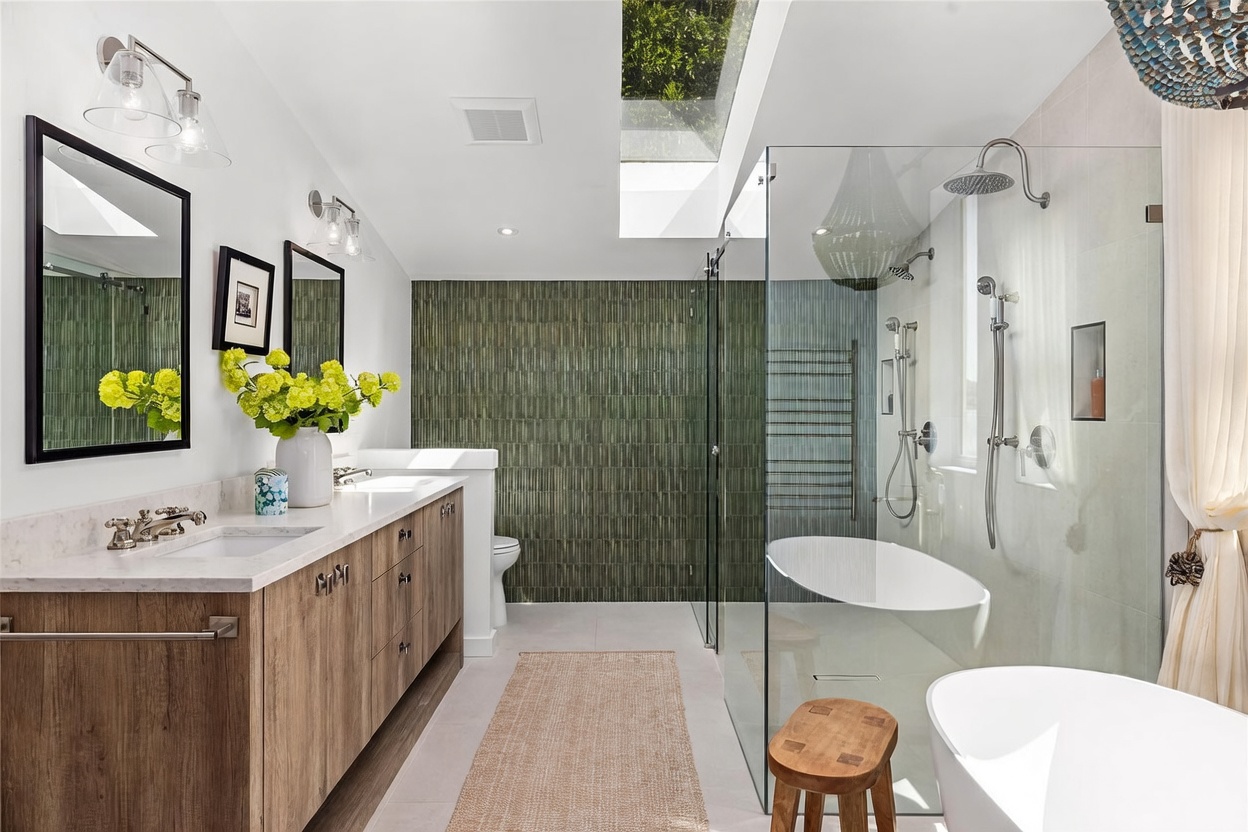 This luxurious primary bathroom features a double vanity with light wood cabinetry and a stone countertop, complemented by a large walk-in shower with a glass enclosure and a separate soaking tub. A striking green textured tile wall serves as a focal point, while a skylight above the shower area floods the space with natural light. The room is finished with a neutral color palette and a small wooden stool, creating a serene and spa-like atmosphere.