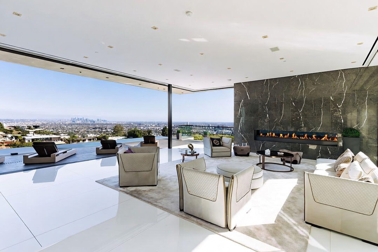 This expansive, ultra-modern living room features a seamless indoor-outdoor flow with floor-to-ceiling glass walls that open to a panoramic city view. The space is anchored by a striking, dark marble fireplace wall and furnished with sophisticated, neutral-toned seating arrangements on a plush area rug. The polished white flooring and minimalist ceiling design create a luxurious, airy atmosphere perfect for high-end entertaining.