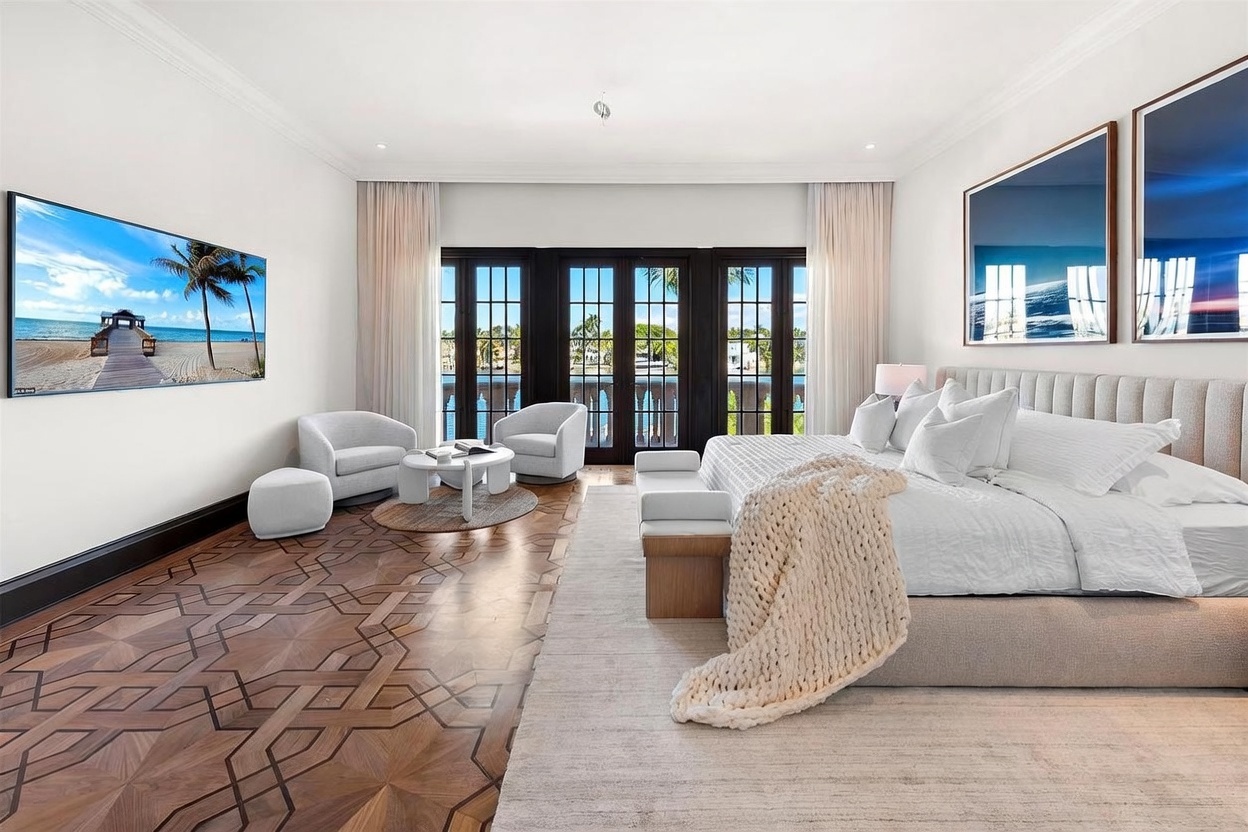 This spacious primary bedroom features intricate patterned wood flooring and a large, plush bed centered against a wall adorned with two large framed artworks. A cozy seating area with two armchairs and a small table sits near the tall glass doors that open to a balcony, while a large wall-mounted television displays a scenic beach view. The room is bathed in natural light, creating an airy and luxurious atmosphere with a neutral color palette.