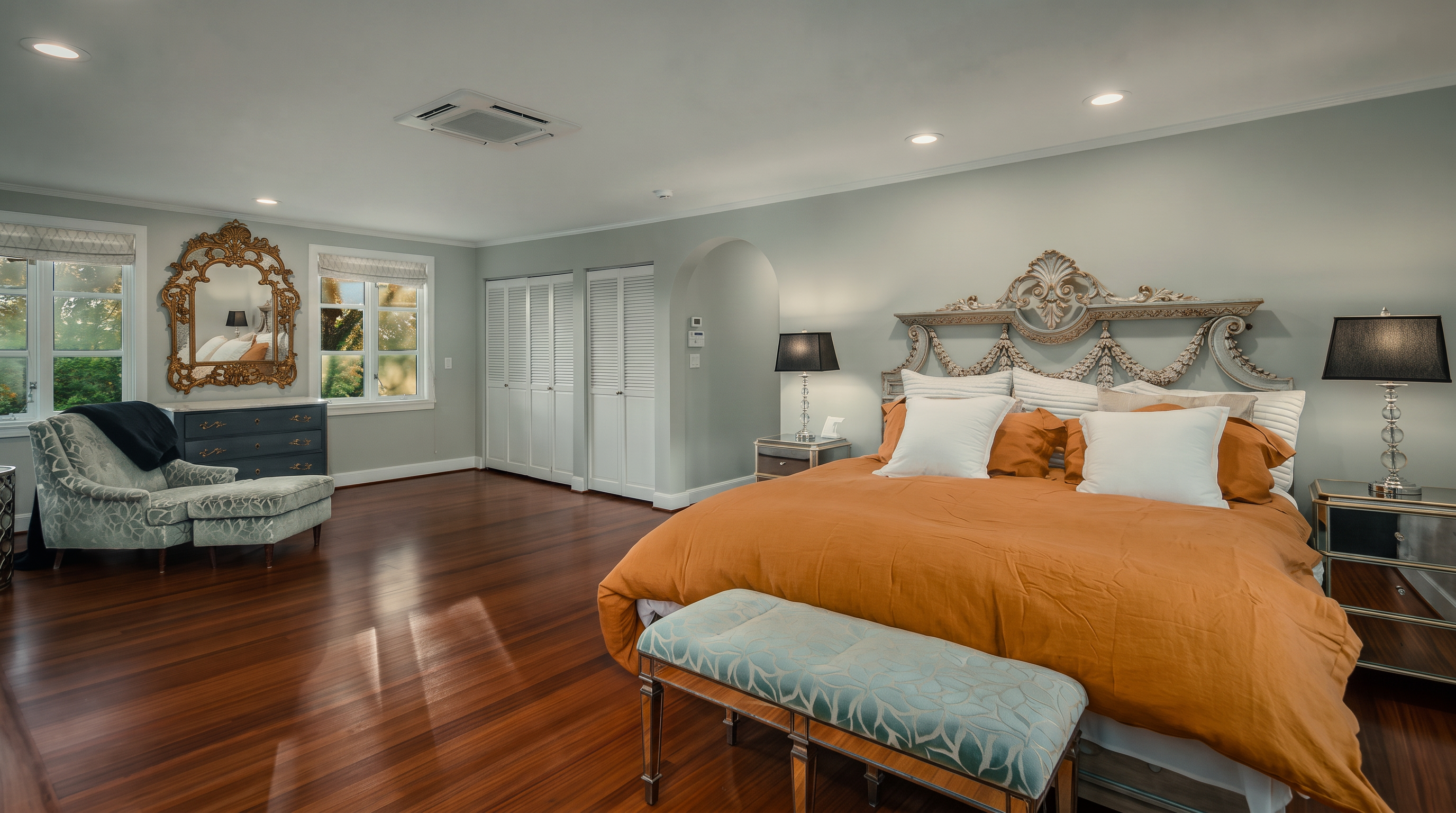This spacious primary bedroom features rich, polished wood flooring and a large, ornate headboard as the focal point. The room is furnished with a plush bed topped with a vibrant orange duvet, a patterned bench at the foot, and a classic chaise lounge near the windows. The neutral wall color and recessed lighting create a bright, inviting atmosphere, while an arched doorway leads to an adjacent space.