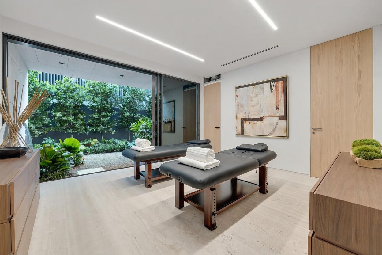 This serene spa or massage room features two professional massage tables centered in the space, each topped with neatly folded white towels. The room is characterized by a minimalist aesthetic, featuring light-toned stone flooring, smooth white walls, and a large abstract painting. A wall of floor-to-ceiling glass sliding doors opens directly onto a lush, private garden, creating a seamless connection between the indoor treatment area and the tranquil outdoor greenery.
