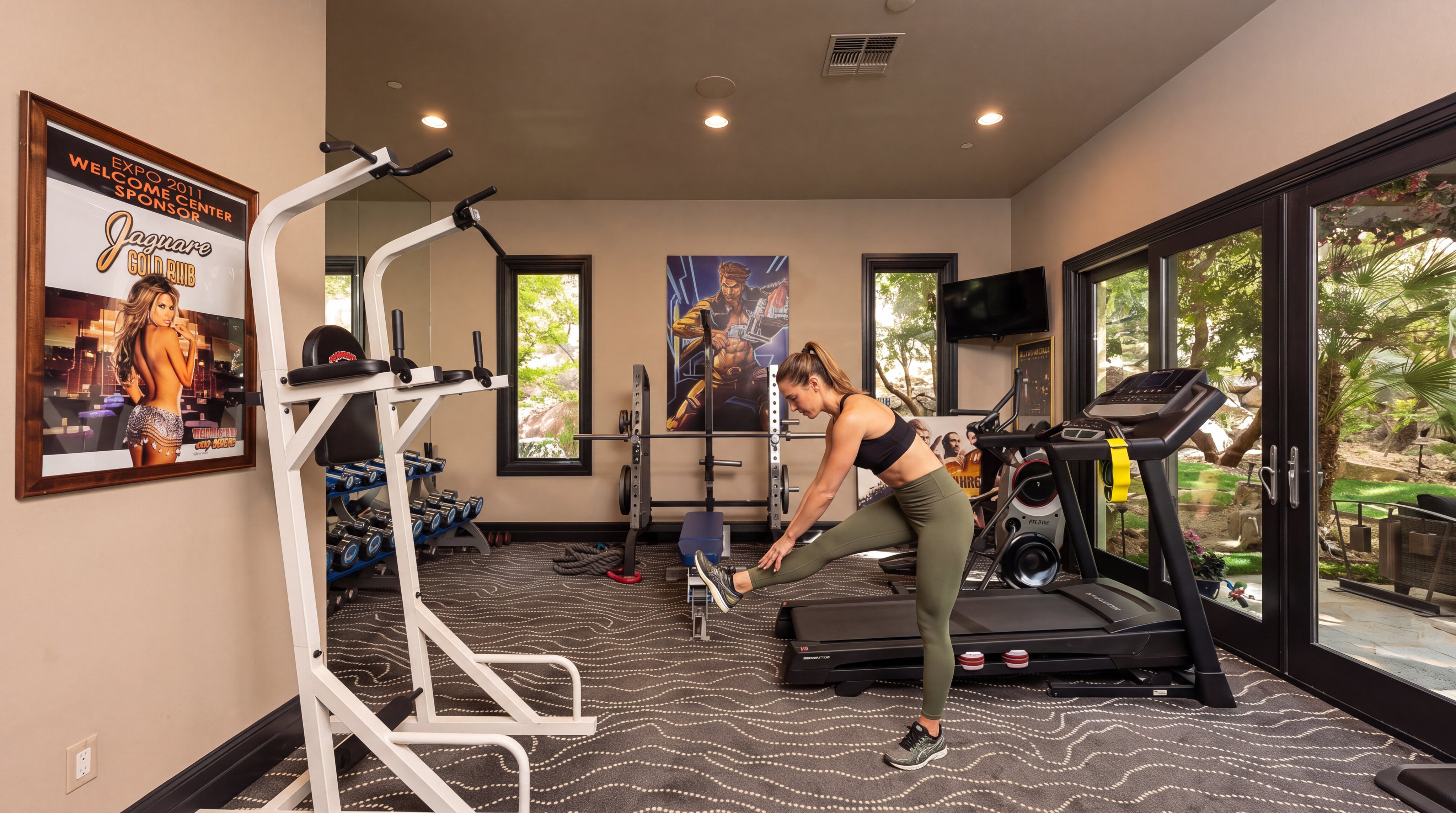 This private home gym is well-equipped with a power rack, treadmill, and free weights, all set against a neutral color palette with patterned carpeting. The space is brightened by natural light coming through floor-to-ceiling glass doors and windows, creating an inviting environment for fitness. Decorative wall art and mirror paneling add character and a sense of depth to the workout room.