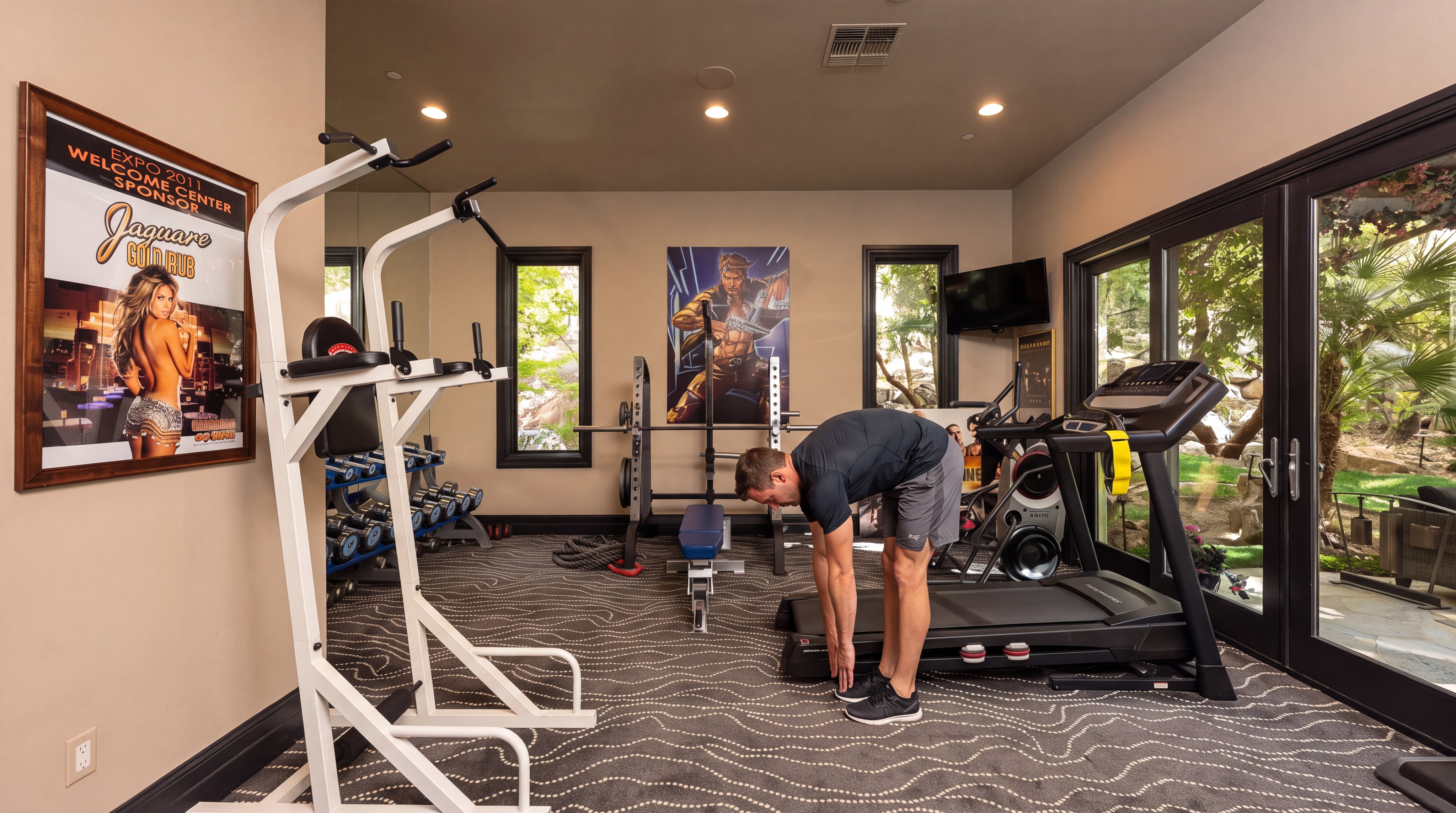 This well-equipped home gym features a variety of workout equipment, including a treadmill, a power rack, and a weight rack with dumbbells. The room includes patterned carpeting, large windows and glass doors that provide natural light, and a personal fitness space with artwork on the walls. It offers a functional and motivational environment for exercise.