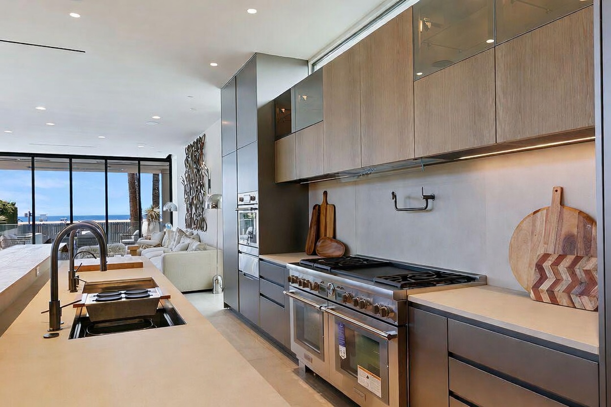 This modern, high-end kitchen features a sleek island with a built-in sink and faucet, paired with dark cabinetry and light wood-toned upper cabinets. A professional-grade stainless steel range and oven are integrated into the cabinetry, while a large circular wooden cutting board rests on the stone countertop. The perspective looks past the kitchen toward an open-concept living area with large glass doors that reveal a bright, coastal view.