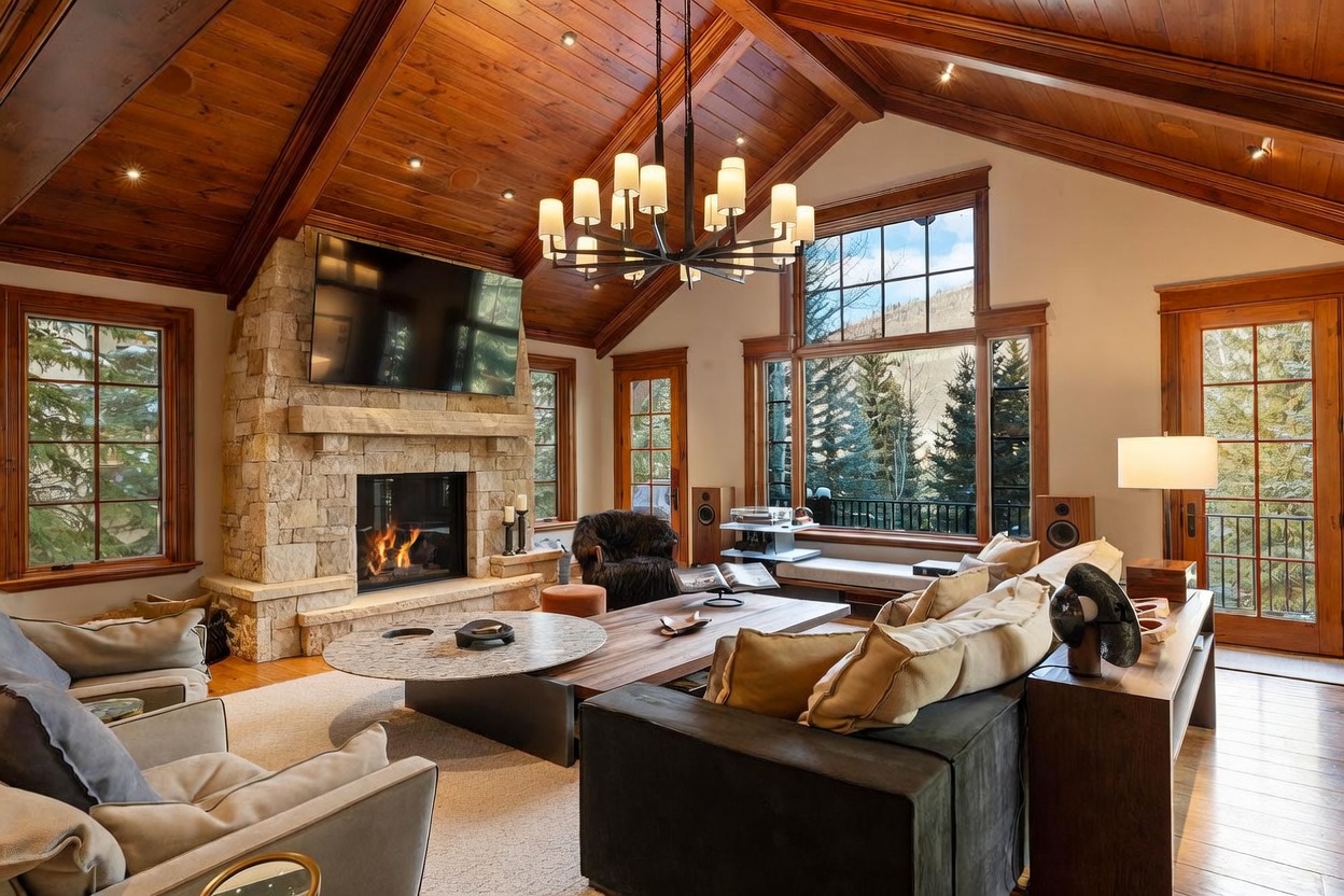 This spacious living room features a vaulted wood-paneled ceiling and a prominent stone-clad chimney breast housing a fireplace. The room is furnished with a large sectional sofa, a round stone-topped coffee table, and a wooden coffee table, all arranged to face the fireplace and the expansive windows that frame a scenic mountain view. A large, multi-armed chandelier serves as a focal point, while the warm wood tones and natural textures create a cozy, mountain-lodge atmosphere.