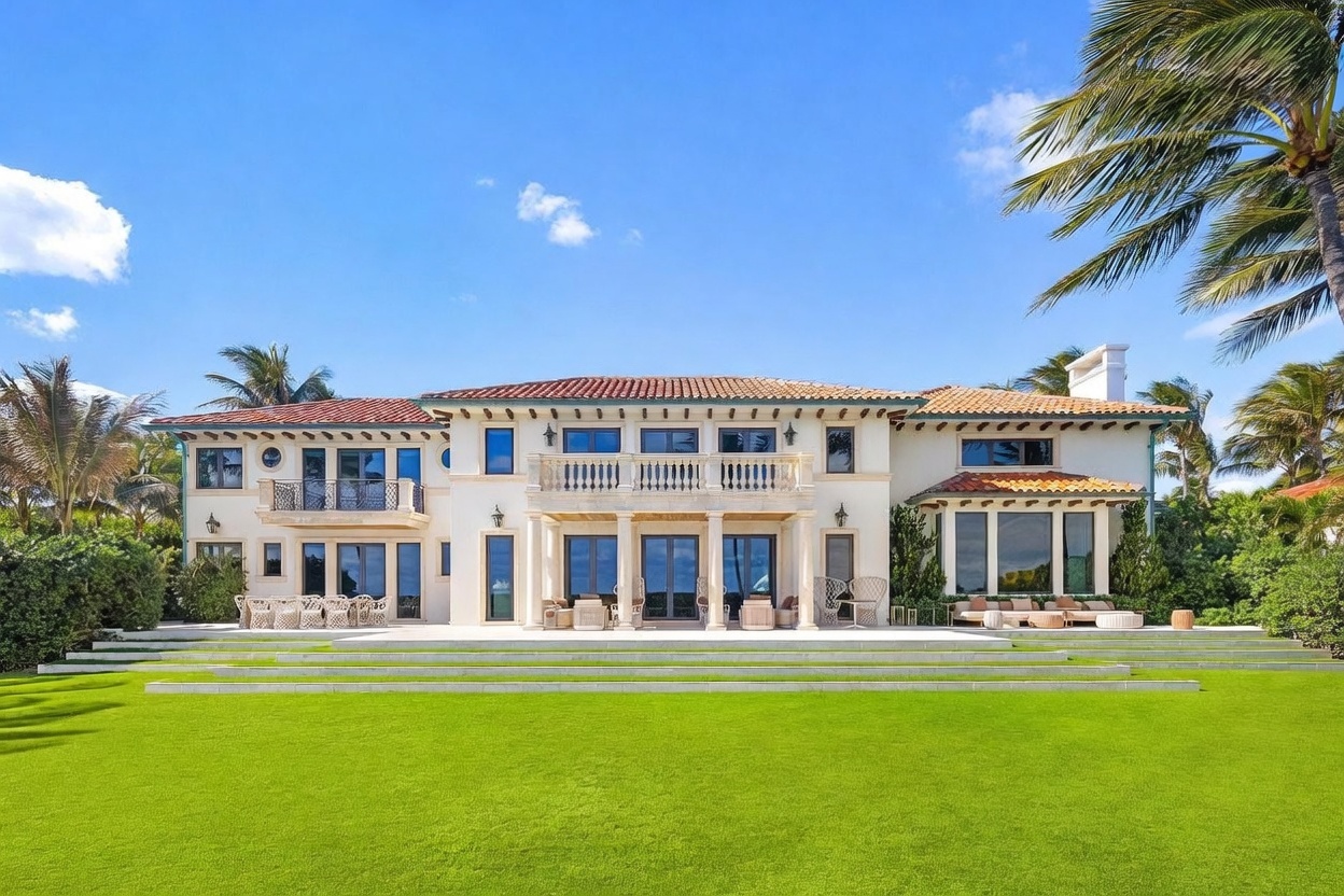 This grand Mediterranean-style estate is captured from a rear perspective, showcasing a symmetrical facade with a terracotta tile roof and light-colored stucco walls. The home features multiple levels of balconies, large windows, and a spacious stone patio furnished with outdoor seating, all overlooking a meticulously manicured lawn. Lush palm trees frame the property against a bright blue sky, emphasizing the luxurious and serene coastal atmosphere.