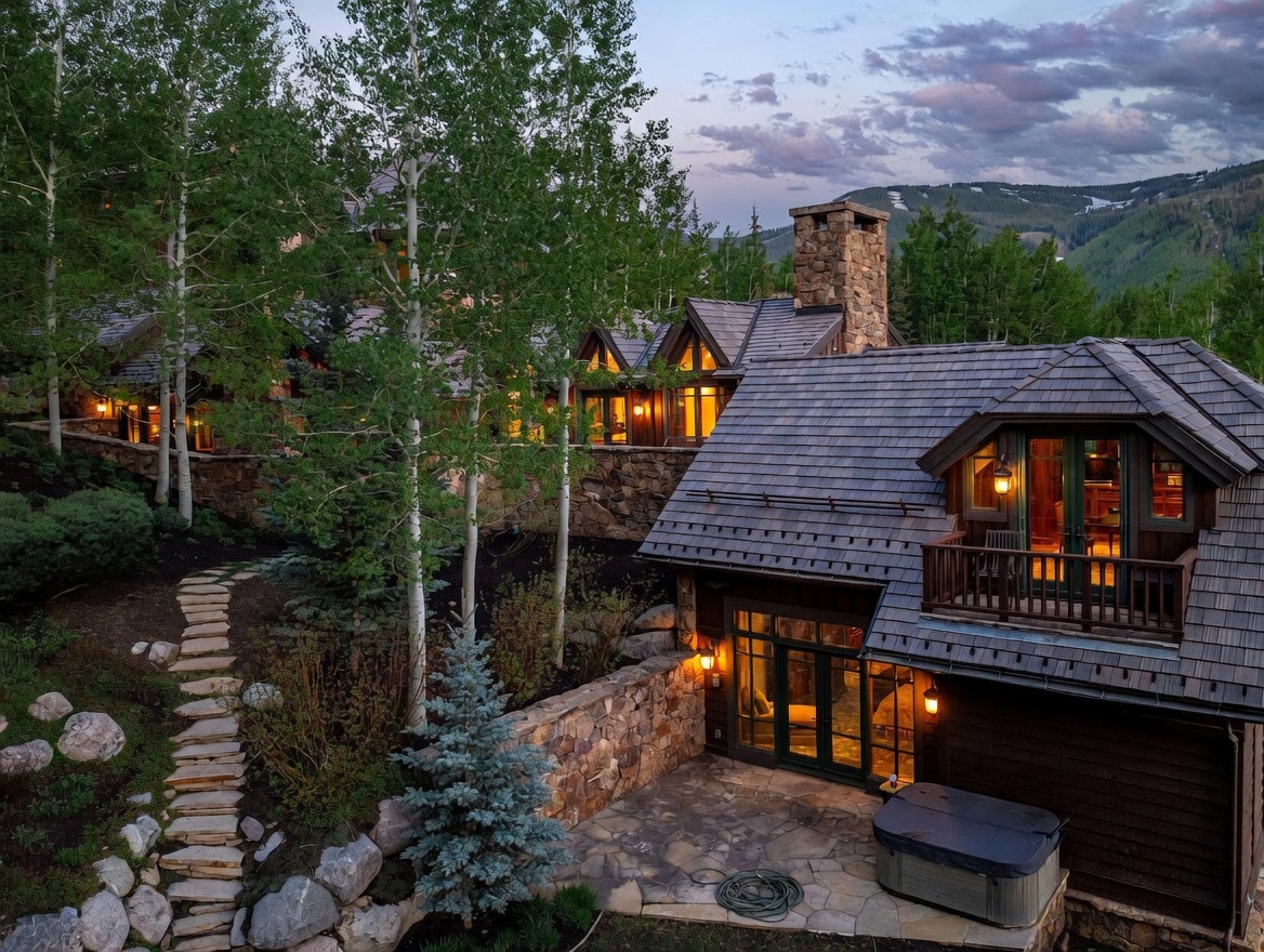 This elevated perspective captures the rear of a rustic mountain estate at dusk, showcasing a multi-level design with dark wood siding and stone accents. The scene features a stone-paved patio equipped with a hot tub, a second-story balcony, and a winding stone pathway leading through a wooded landscape. The warm glow from the interior lighting contrasts beautifully with the twilight sky and the surrounding aspen trees.