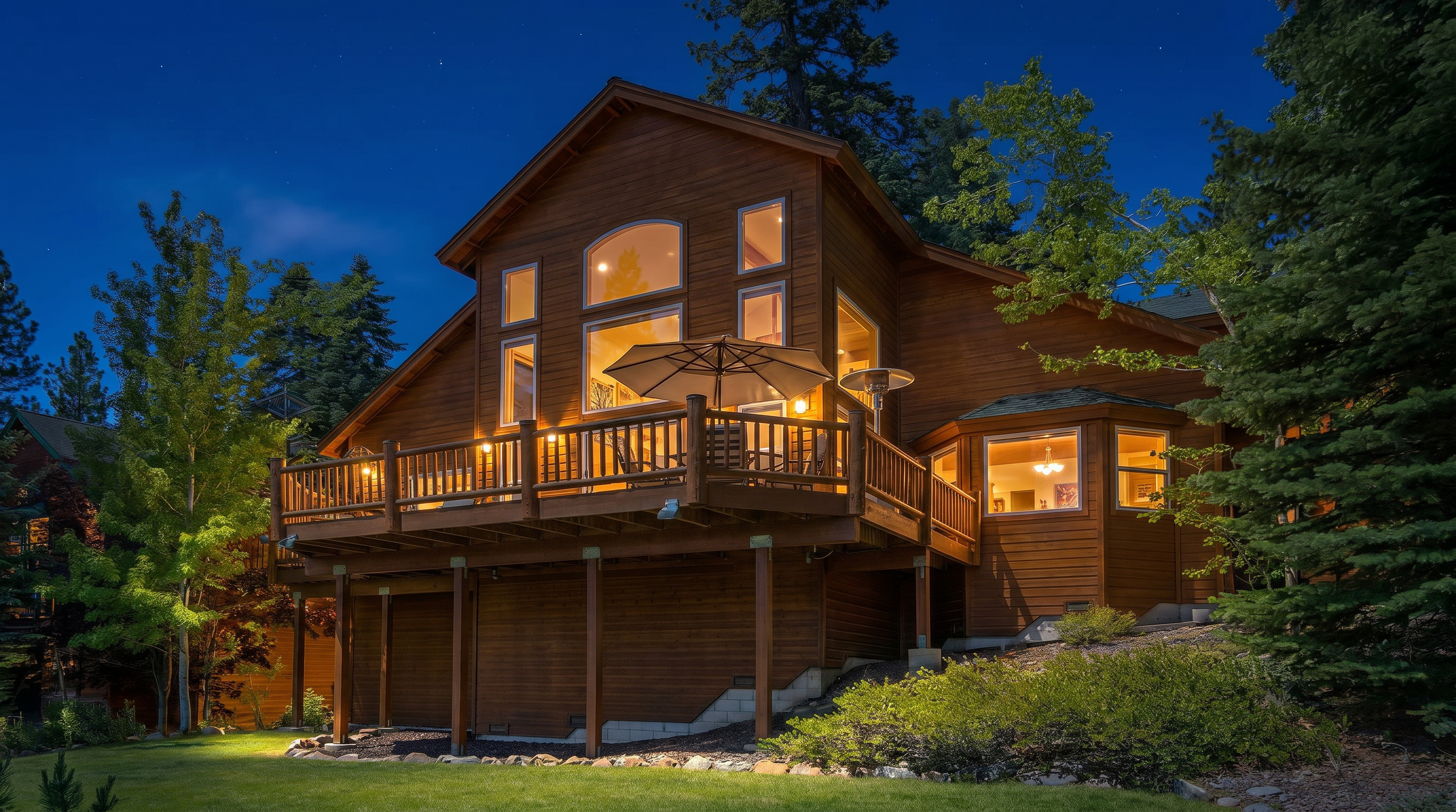 This stunning mountain-style cabin is captured at dusk, showcasing a large, elevated wrap-around wooden deck that perfectly bridges the indoor and outdoor living spaces. The exterior features warm-toned wood siding, expansive multi-level windows that glow with inviting interior light, and a picturesque forest setting. The home exudes a cozy, rustic luxury aesthetic, ideal for evening relaxation and entertaining.