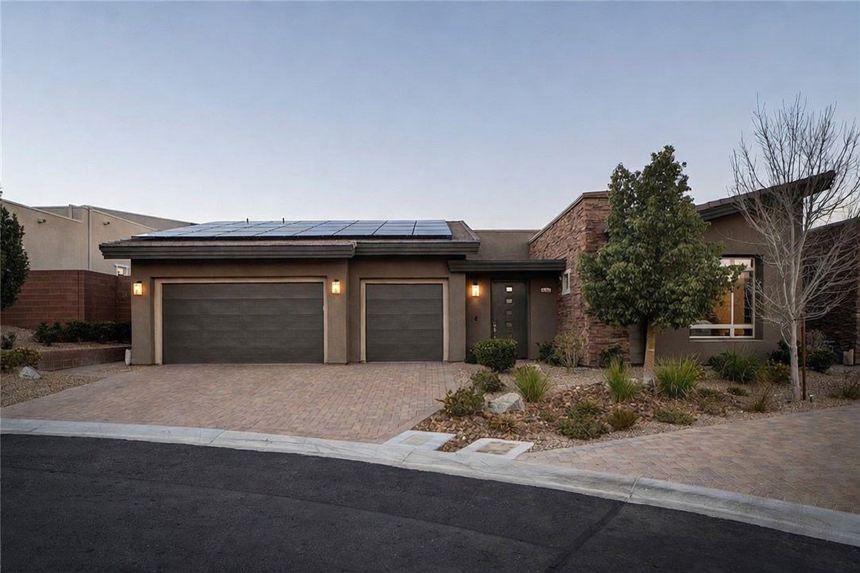 This modern single-story residence features a contemporary architectural design with a combination of smooth stucco and textured stone cladding. The front facade includes a two-car garage, a recessed entryway, and a low-profile roof equipped with solar panels. A paved driveway leads to the home, complemented by desert-inspired landscaping with small shrubs and decorative gravel.