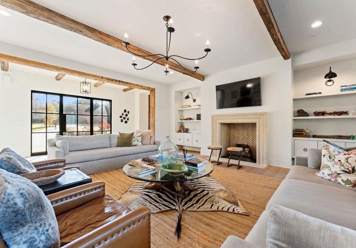 This spacious living room features a warm, rustic aesthetic with exposed wooden ceiling beams and a central stone fireplace surround. The room is furnished with a large light-colored sofa, leather armchairs, and a circular glass-topped coffee table resting on a patterned animal-print rug. Large, grid-paned windows provide a view of the outdoors, while built-in shelving units flank the fireplace, creating a balanced and inviting atmosphere.