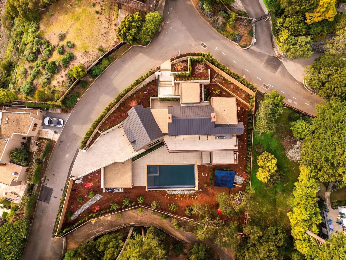 This high-angle aerial perspective captures a modern, multi-level residence nestled on a landscaped hillside lot. The property features a sleek, dark-roofed structure, a rectangular swimming pool, and expansive paved patio areas surrounded by terraced gardens and mature trees. The home is situated at a corner intersection, showcasing its unique architectural footprint and integration into the sloped terrain.