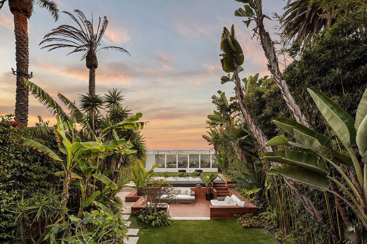 This lush, tropical-inspired backyard features a multi-level wooden deck with built-in seating, leading to a rectangular swimming pool. The property is framed by dense, mature greenery and tall palm trees, creating a private oasis that overlooks the ocean in the distance. The scene is captured at sunset, casting a warm, golden glow over the entire outdoor living space.
