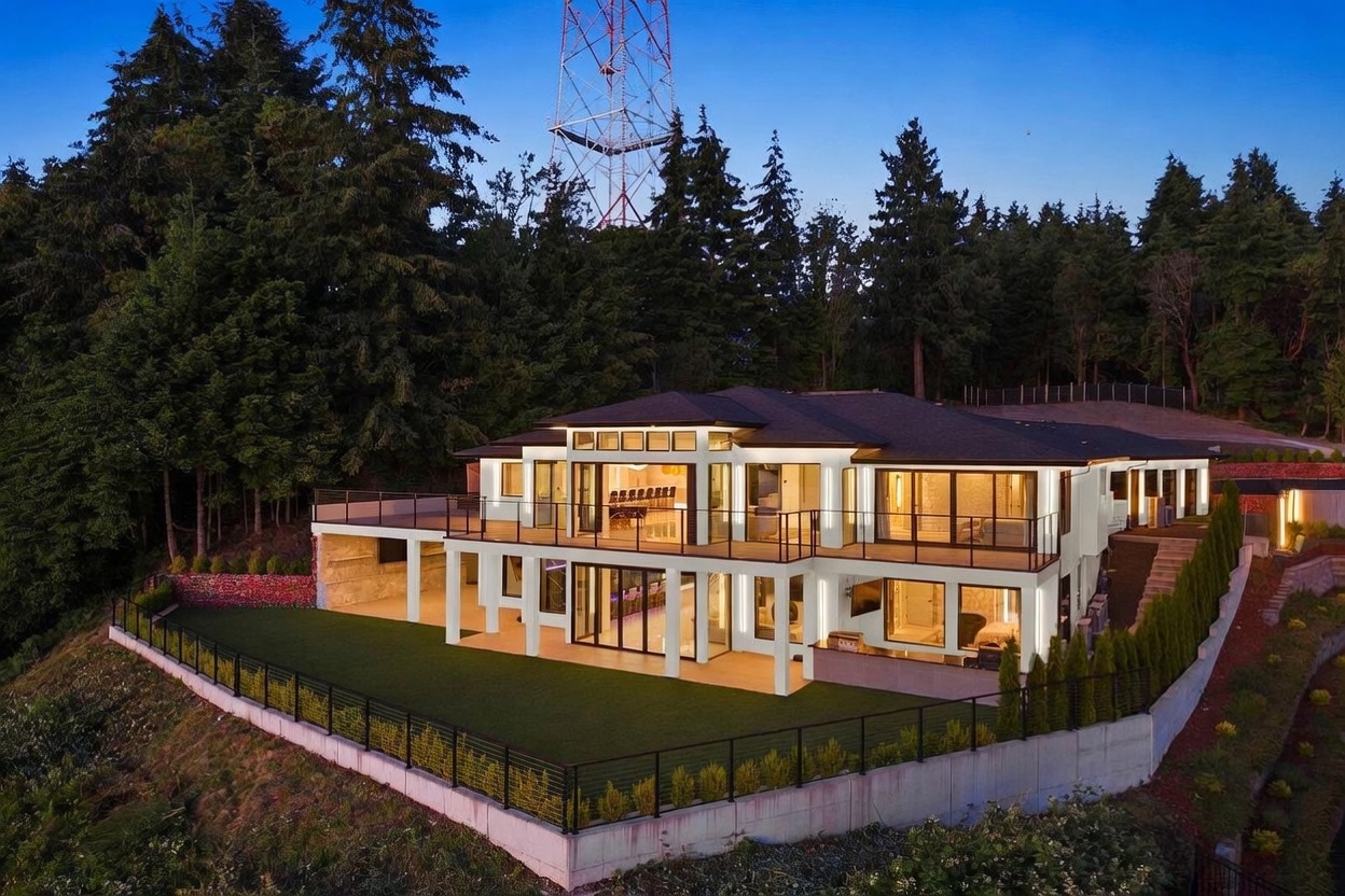 This aerial perspective captures a modern, multi-level luxury residence set against a backdrop of dense forest and a prominent communications tower. The home features a clean, contemporary design with expansive glass walls, a spacious upper deck, and a lower-level patio that opens onto a manicured lawn enclosed by a concrete retaining wall. The warm interior lighting contrasts beautifully with the twilight sky, highlighting the home's architectural lines and seamless indoor-outdoor living spaces.