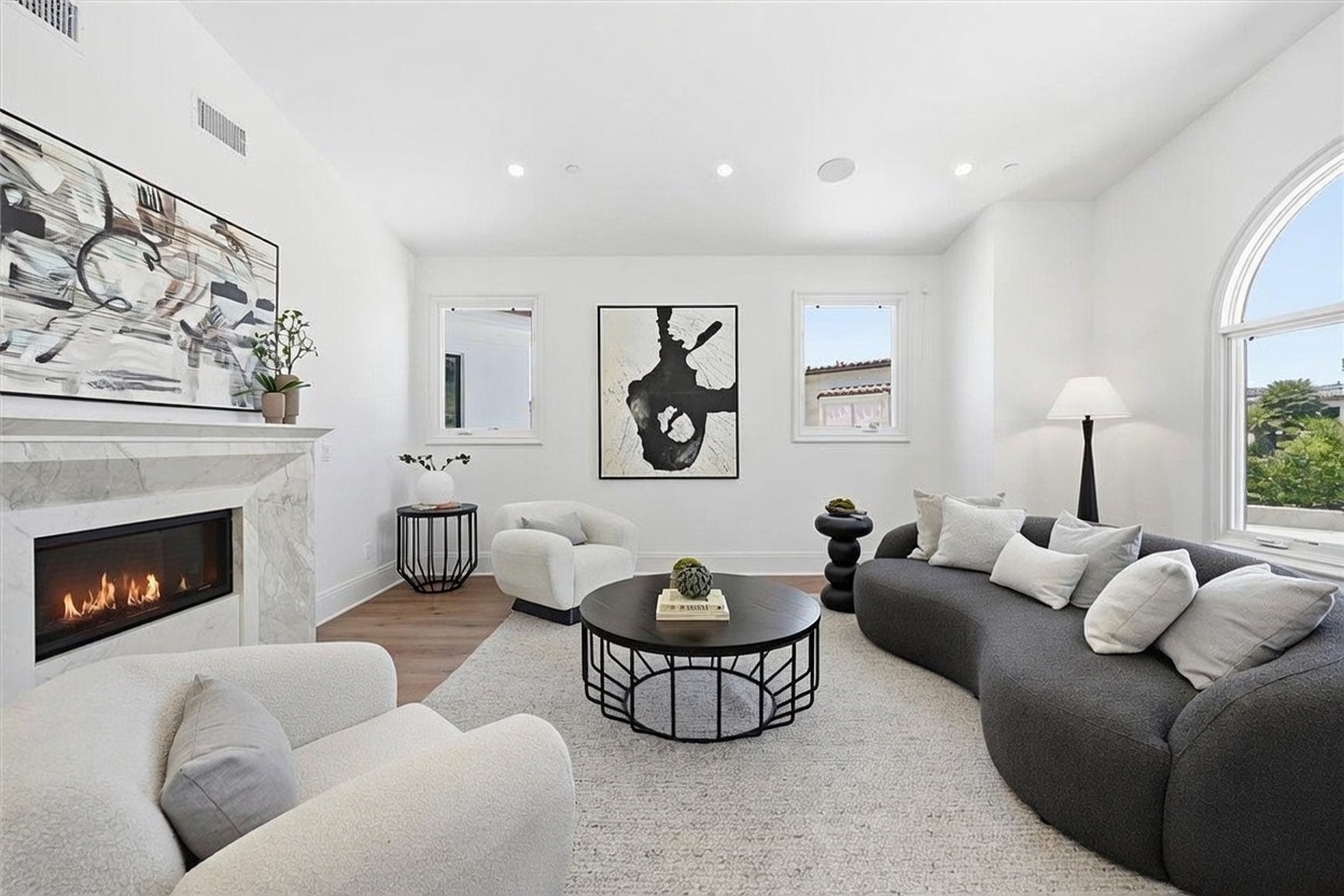 This bright and airy living room features a contemporary design with a curved dark gray sofa, two cream-colored armchairs, and a round black coffee table. A fireplace with a stone surround serves as a focal point on the left, while large arched windows on the right allow for ample natural light. The space is accented by modern abstract artwork on the walls and a neutral area rug, creating a sophisticated and inviting atmosphere.