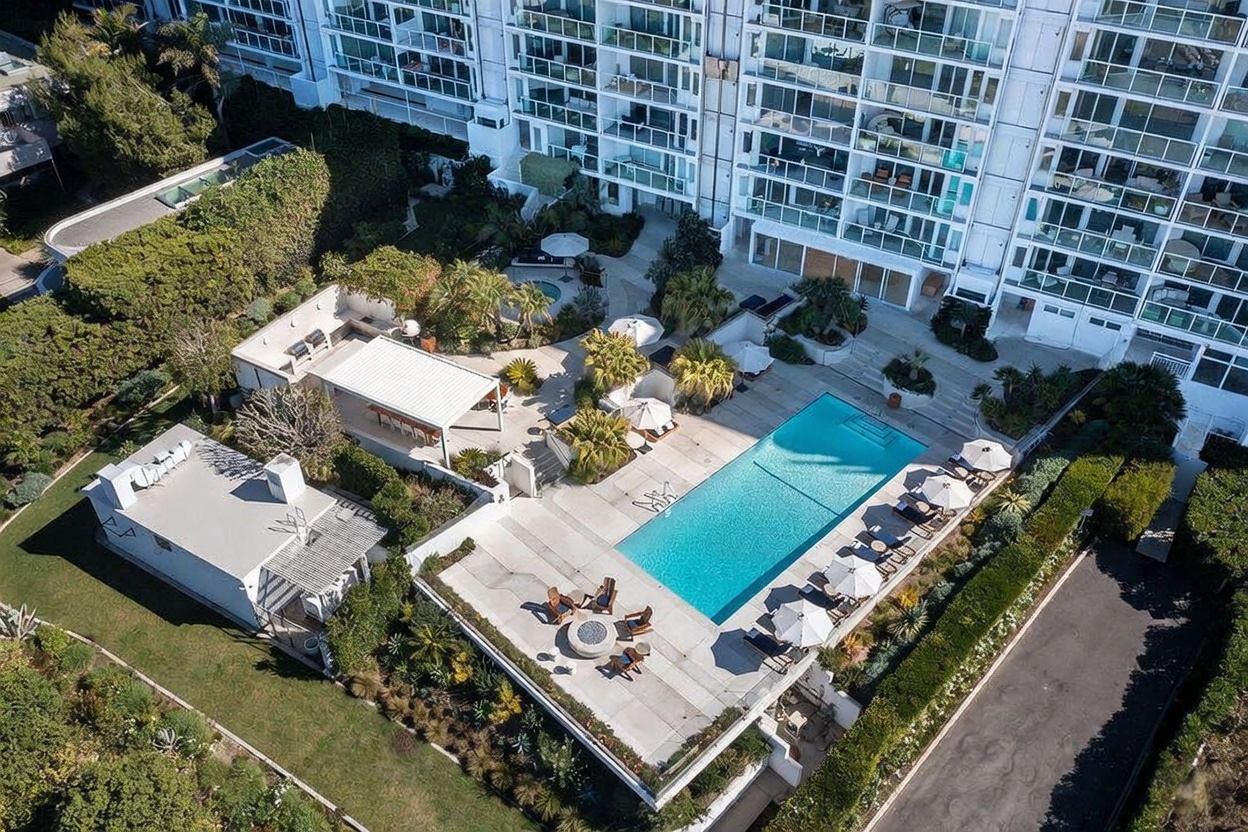 This high-angle aerial view captures a luxury residential complex featuring a large, rectangular swimming pool surrounded by a spacious concrete deck. The outdoor area includes a covered lounge space, a fire pit seating area, and lush landscaping, all situated adjacent to a multi-story building with numerous balconies. The perspective provides a comprehensive look at the property's recreational amenities and its integration with the surrounding greenery.