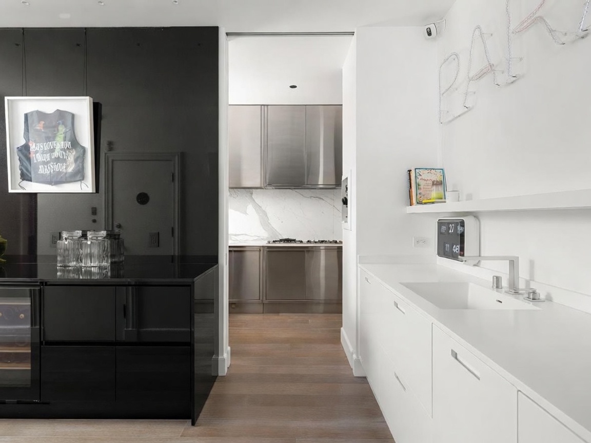 This modern kitchen space features a striking contrast between a glossy black island and cabinetry on the left and a clean white countertop and cabinetry area on the right. A central doorway leads into a secondary kitchen area with metallic-finish cabinets and a stone backsplash. The room is accented by contemporary art, a digital clock, and a neon sign, creating a sleek and sophisticated aesthetic.