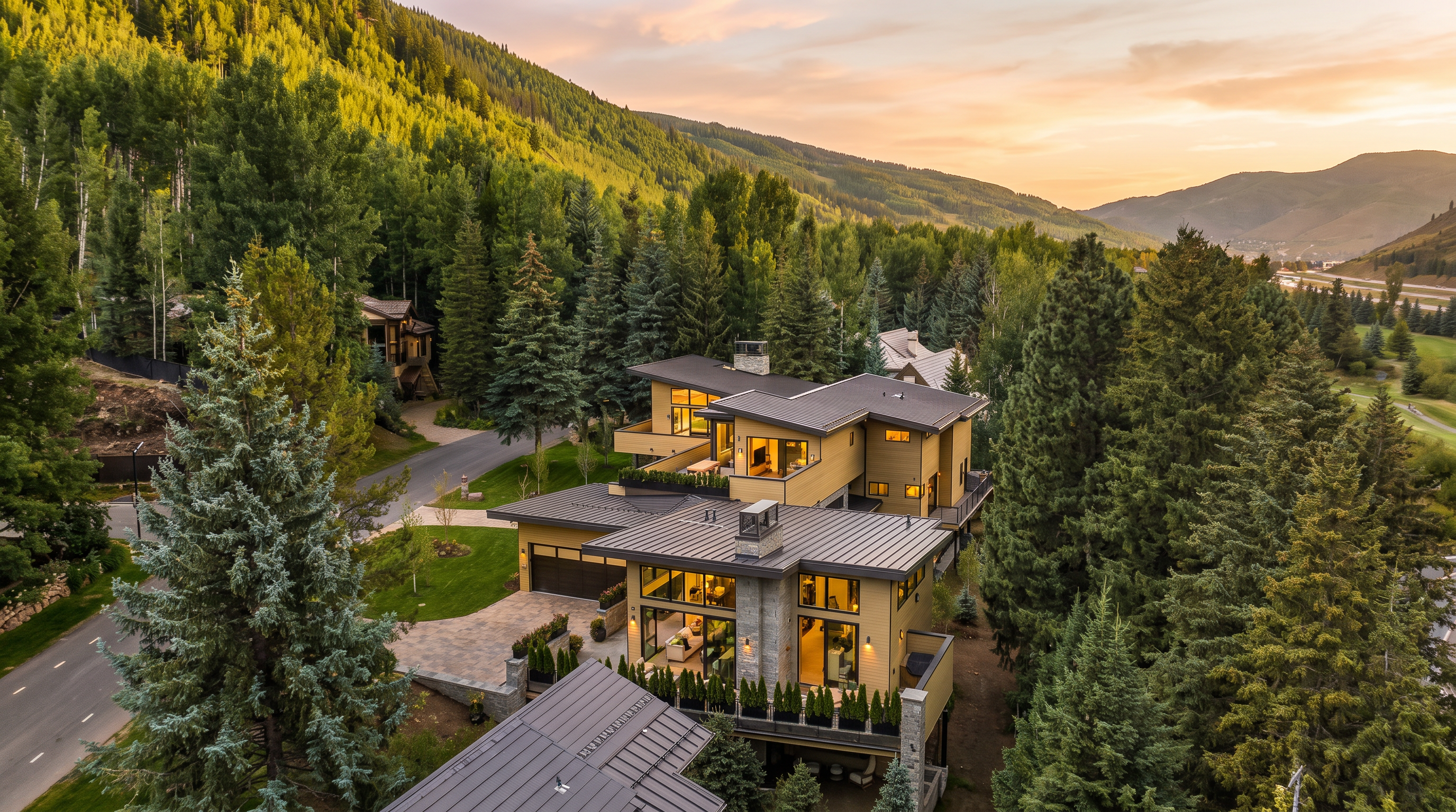 This high-angle aerial view captures a modern, multi-level mountain residence nestled within a lush, forested landscape. The home features a contemporary design with dark metal roofing, light-toned siding, and expansive glass walls that offer seamless indoor-outdoor transitions. Surrounded by dense pine trees and a winding road, the property exudes a sense of secluded luxury and architectural sophistication.