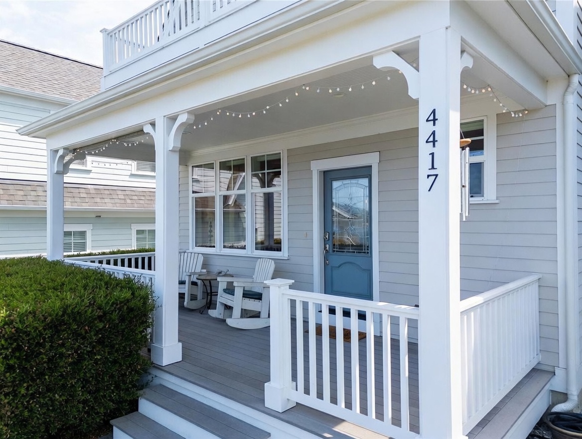 This inviting front porch features light gray horizontal siding, a blue front door with decorative glass, and a set of white rocking chairs. The space is accented with string lights hanging from the porch ceiling and a white railing that encloses the deck. The perspective is a low-angle, eye-level shot that highlights the welcoming curb appeal and the architectural details of the home's entrance.