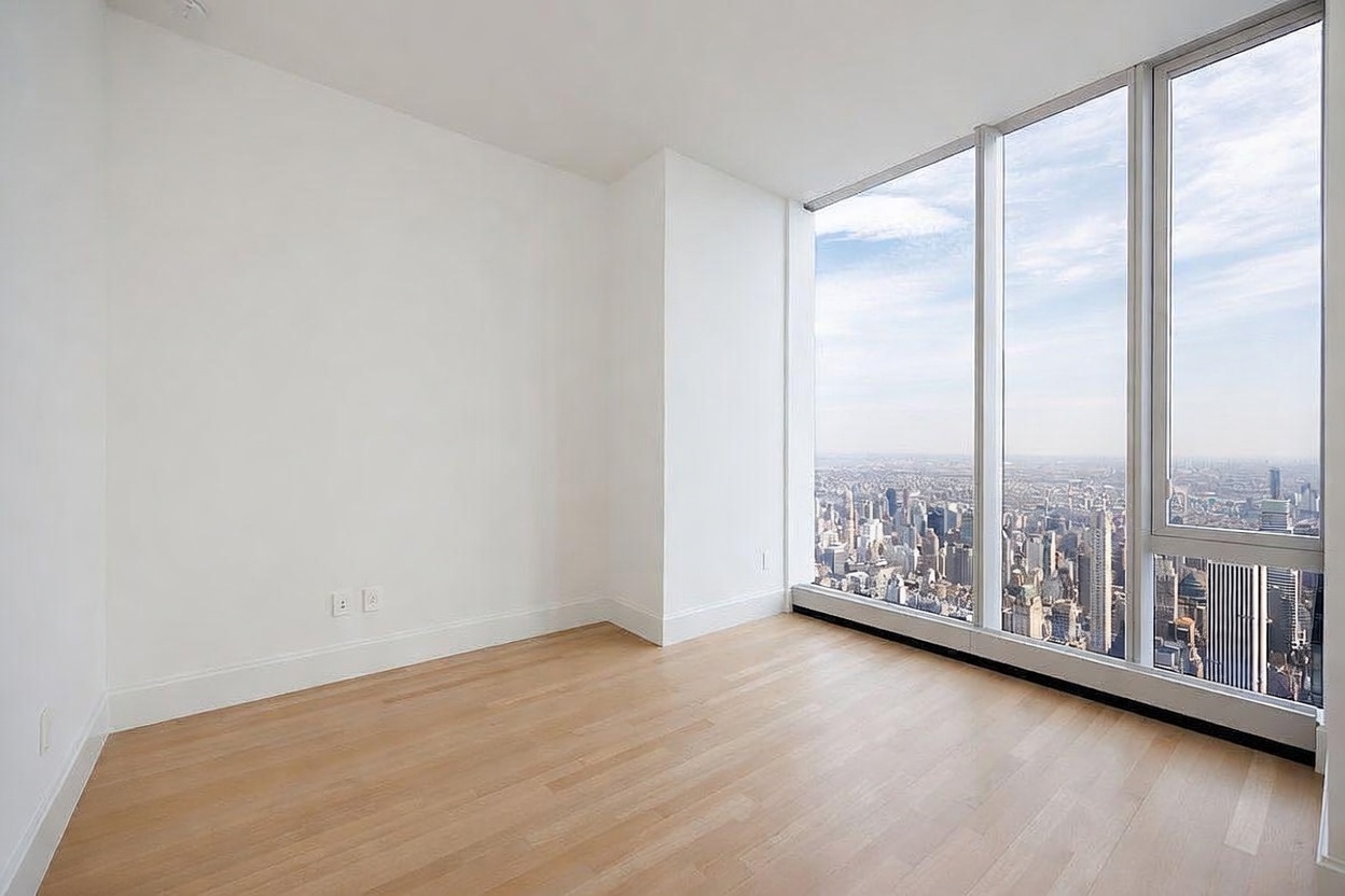 This minimalist bedroom features light wood flooring and crisp white walls, creating a bright and airy atmosphere. The space is defined by expansive floor-to-ceiling windows that provide a dramatic, unobstructed panoramic view of the city skyline. The room's clean lines and neutral palette emphasize the impressive height and urban perspective.