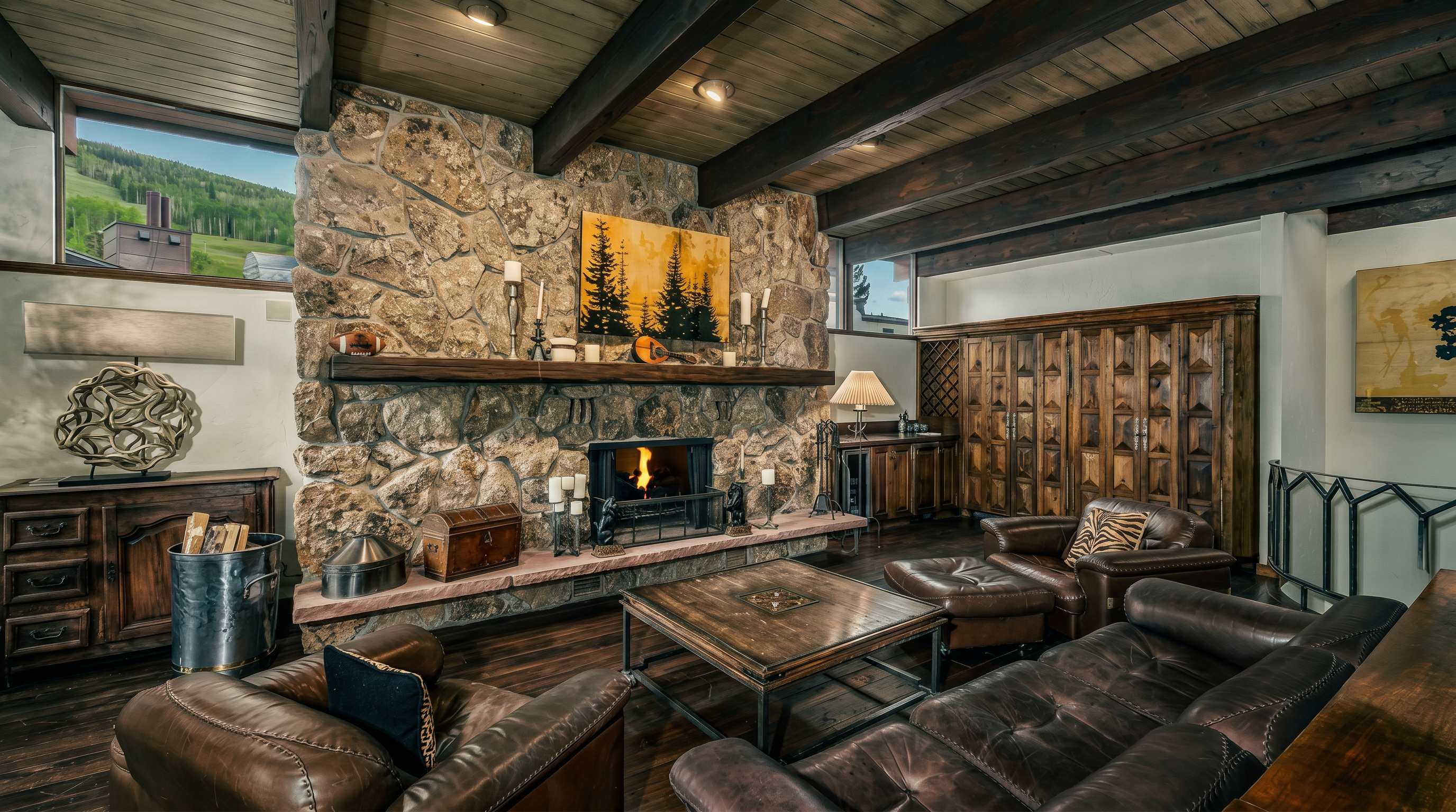 This rustic living room features a prominent stone-clad chimney breast with a heavy timber mantel and a crackling fire. The space is furnished with rich brown leather seating, a large wooden coffee table, and exposed wooden ceiling beams that add to the mountain-lodge aesthetic. Large windows flanking the stone feature provide natural light and views of the surrounding landscape, while a decorative wooden cabinet wall adds warmth and storage.