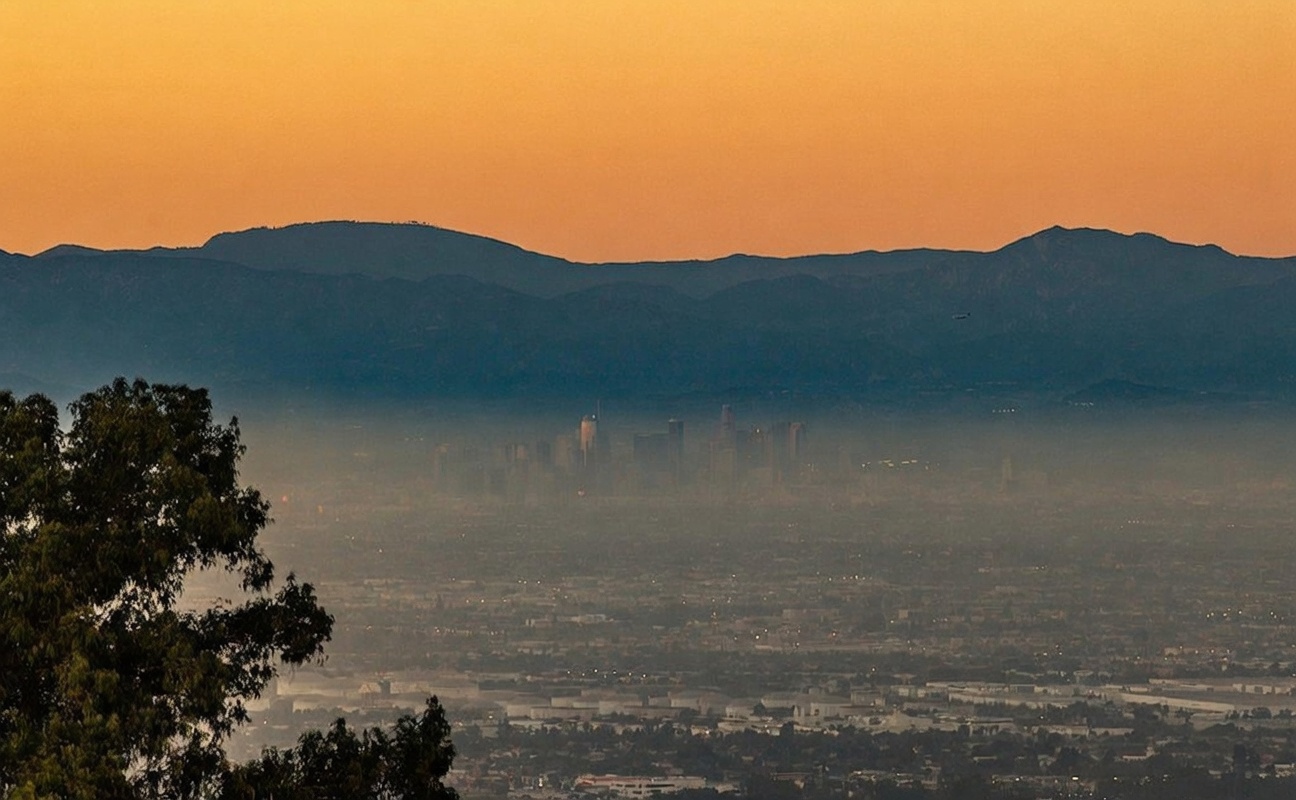 This scenic landscape shot captures a panoramic view of a city skyline nestled in a valley beneath a hazy, golden sunset. In the foreground, the dark silhouettes of tree branches frame the vista, while the distant mountains provide a dramatic backdrop. The perspective is elevated, suggesting a high-vantage point from a hillside property overlooking the urban sprawl.