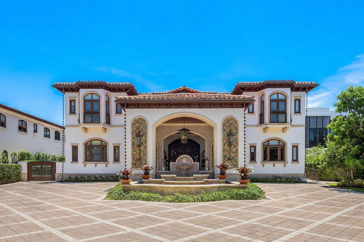 This grand Mediterranean-style estate features a symmetrical facade with a prominent arched entryway, decorative tile accents, and a central stone fountain. The expansive paved driveway leads directly to the impressive front entrance, framed by lush landscaping and mature trees. The architecture exudes luxury and elegance, characterized by its warm stucco walls, dark wood trim, and classic clay tile roof.