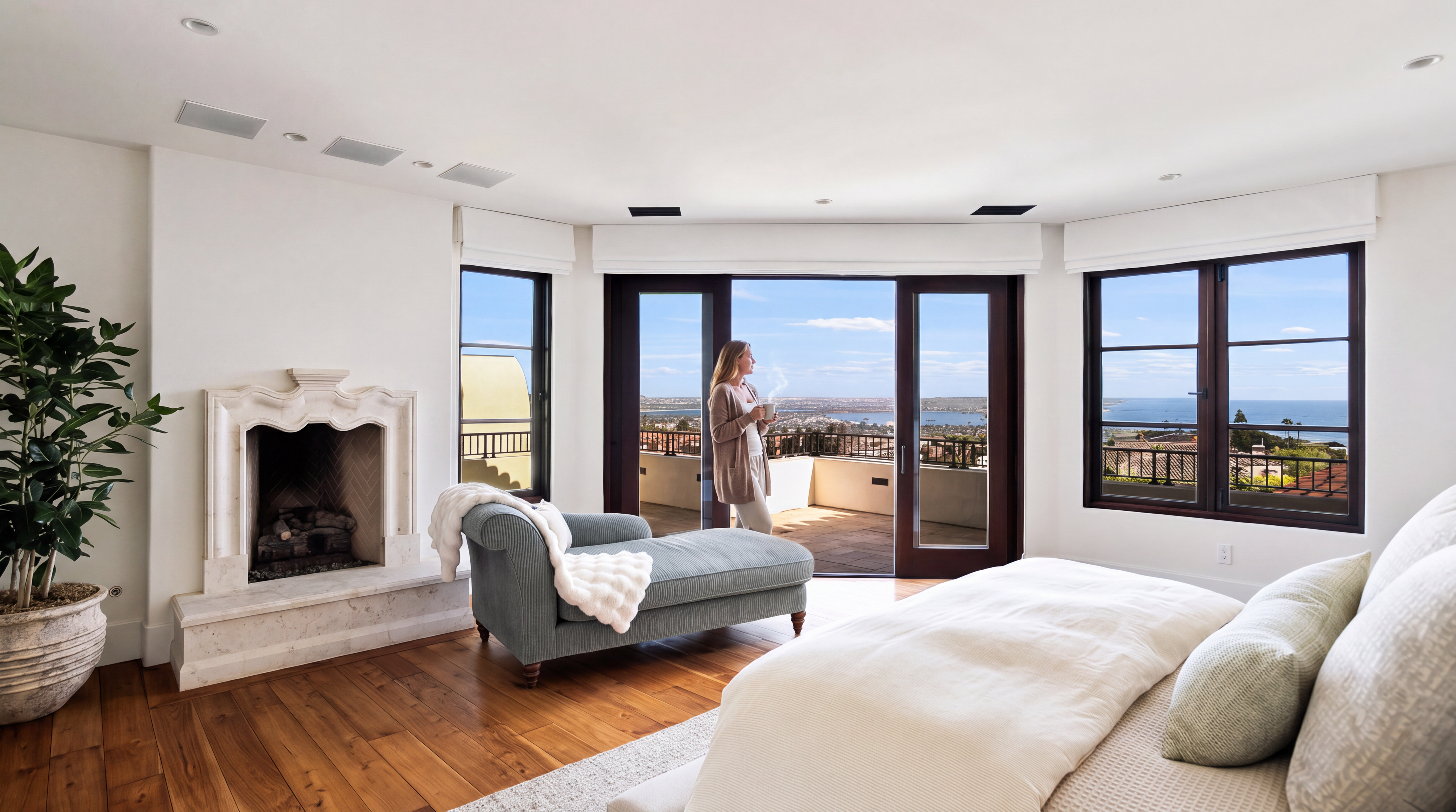 This luxurious primary bedroom features a warm, inviting atmosphere with rich hardwood flooring, an elegant fireplace, and a plush chaise lounge. Expansive sliding glass doors open onto a scenic balcony that offers stunning, elevated panoramic views of the coast and bay. The space is beautifully appointed, blending comfort and sophistication for a serene retreat.
