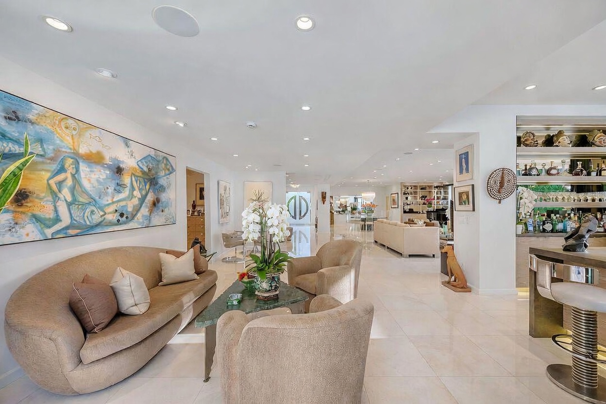 This expansive, open-concept living area features polished light-colored flooring and a sophisticated, neutral color palette. A curved sofa and plush armchairs are arranged around a central stone-topped coffee table, while a large, vibrant abstract painting serves as a focal point on the wall. The space flows seamlessly into a secondary seating area and a bar, creating an airy and luxurious atmosphere suitable for entertaining.