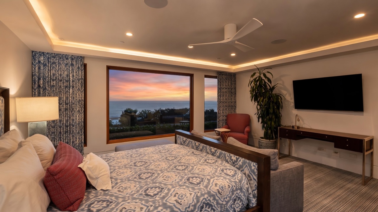 This spacious primary bedroom features a large bed with a patterned blue and white duvet, positioned to face a wall-mounted television and a sleek dark wood console table. Large windows provide a panoramic view of the ocean at dusk, while a cozy red armchair and a tall potted plant add warmth to the corner. The room is illuminated by recessed lighting and a modern ceiling fan, creating a serene and sophisticated atmosphere.