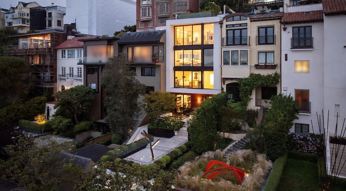 This elevated rear view captures a multi-level urban residence featuring a striking modern glass-fronted addition nestled among traditional neighboring homes. The property boasts a meticulously landscaped terraced garden with stone pathways, lush greenery, and a prominent red sculptural art piece. The perspective highlights the architectural contrast between the contemporary glass facade and the classic multi-story structures, creating a sophisticated and private outdoor living environment.