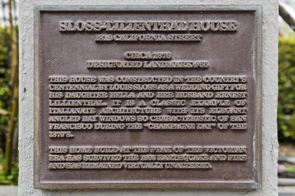 This image features a bronze historical landmark plaque mounted on a textured, light-colored wall. The plaque provides detailed information about the Sloss-Lilienthal House, including its 1876 construction date, architectural style, and historical significance. The close-up, eye-level perspective highlights the embossed lettering and the weathered, metallic texture of the commemorative marker.