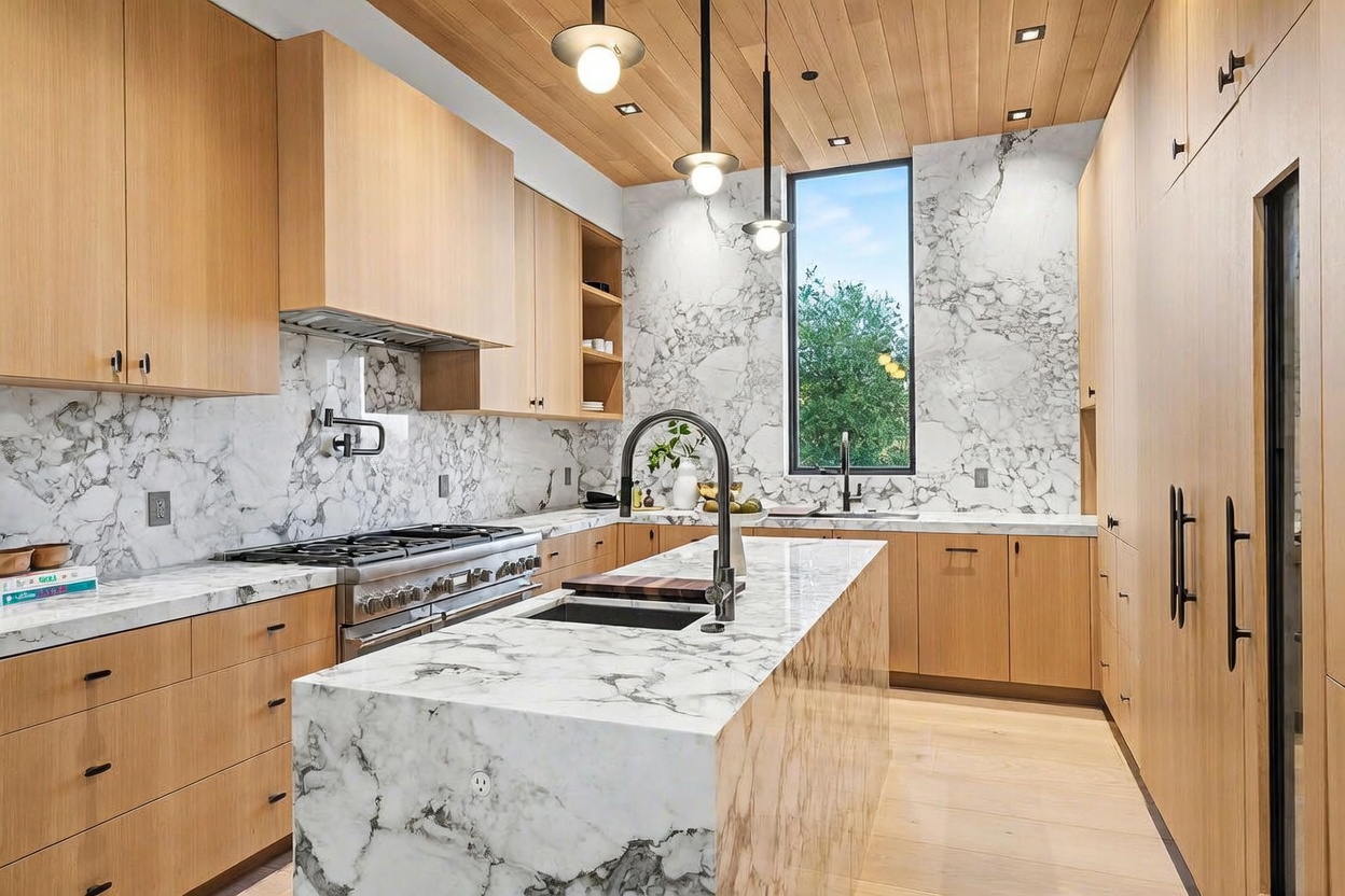 This modern kitchen features light wood cabinetry and a prominent central island topped with patterned stone. The space is accented by a matching stone backsplash, a professional-style range, and three pendant lights hanging from a wood-paneled ceiling. A tall window above the sink area provides natural light, creating a bright and sophisticated atmosphere.