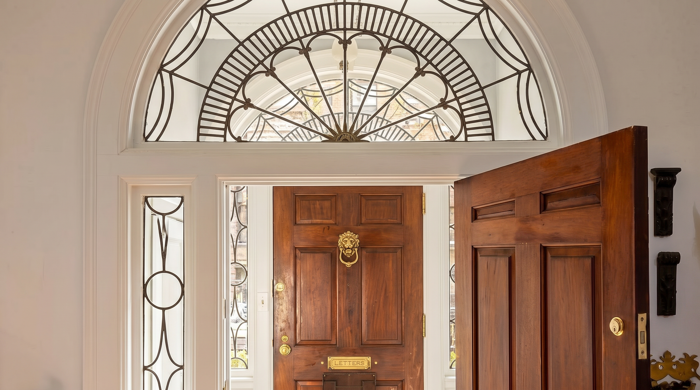 This elegant entryway features a grand, arched doorway with intricate wrought-iron detailing above the transom. The perspective looks through the open, rich-toned wooden front door into a foyer with classic black-and-white checkered marble flooring and a secondary wooden door. The architectural style is sophisticated and historic, emphasizing a welcoming and high-end residential aesthetic.
