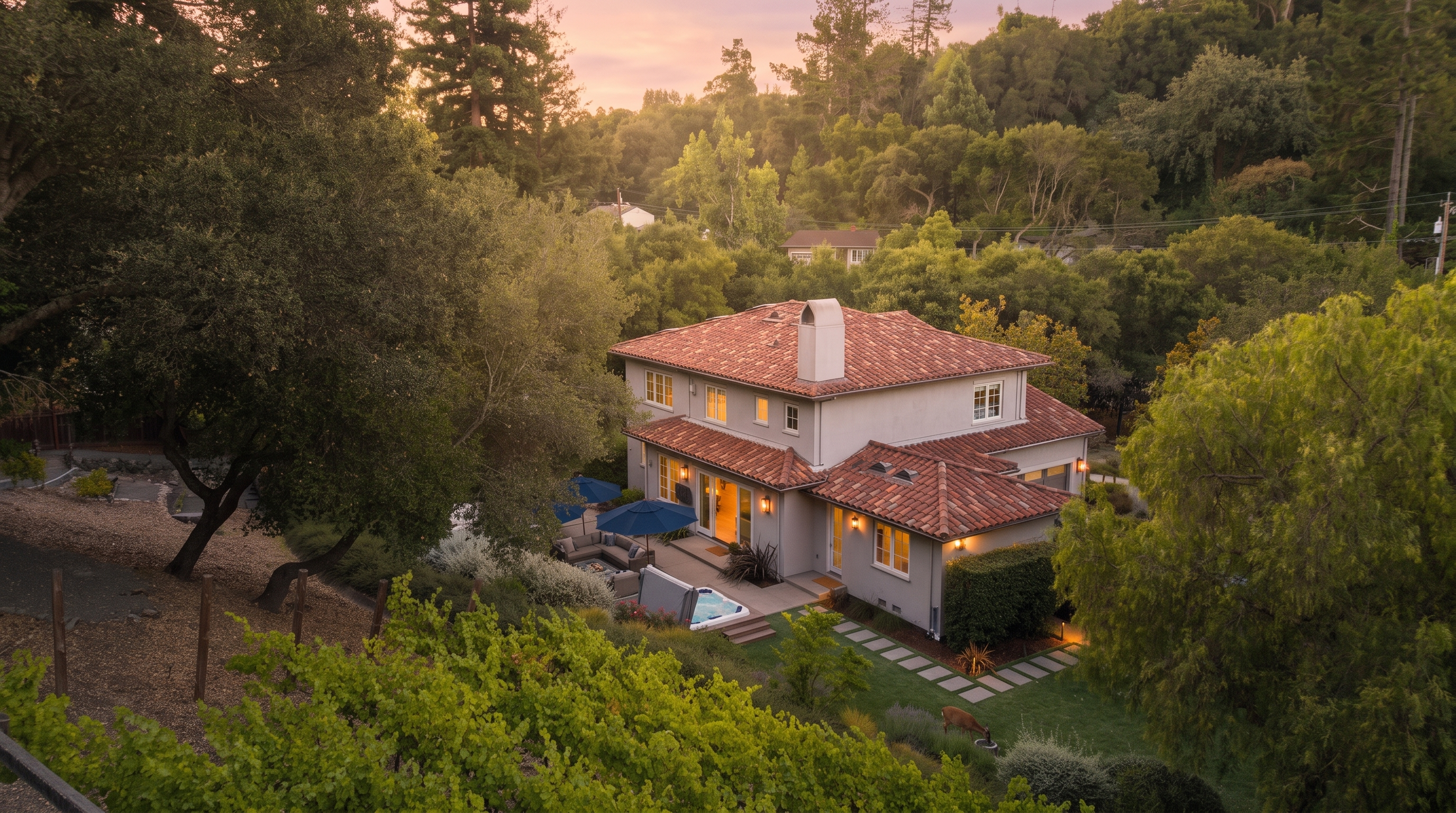This aerial view showcases a charming two-story home with a classic Mediterranean-inspired tile roof, nestled within a lush, wooded landscape. The property features a well-manicured backyard with a patio area, a hot tub, and a small vineyard in the foreground, creating an inviting and serene atmosphere. Its elegant architecture and peaceful natural surroundings make it a standout, high-end residential property.