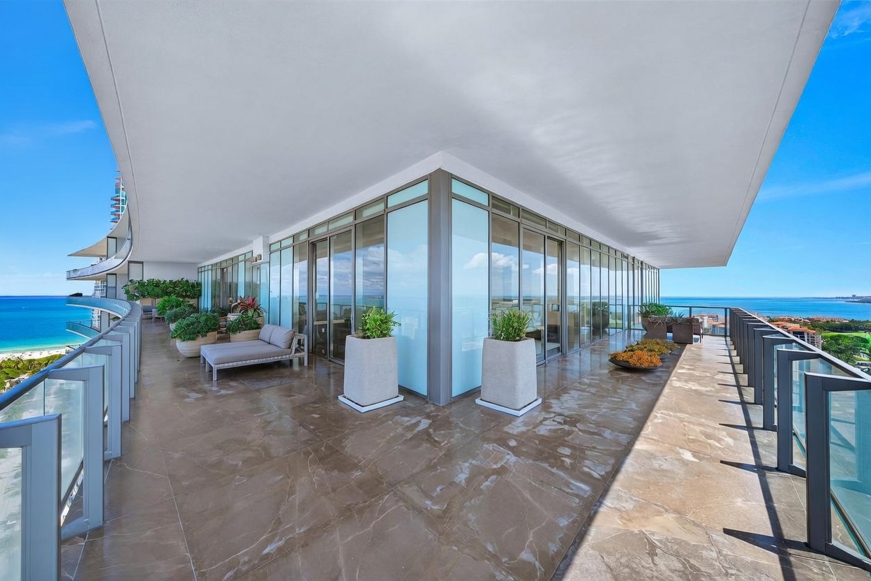 This expansive, wrap-around balcony features polished stone flooring and floor-to-ceiling glass walls that offer panoramic ocean views. The space is furnished with a sleek lounge chair and several large planters, creating a sophisticated outdoor living area. The perspective captures the corner of the building, emphasizing the seamless transition between the interior living space and the vast coastal horizon.