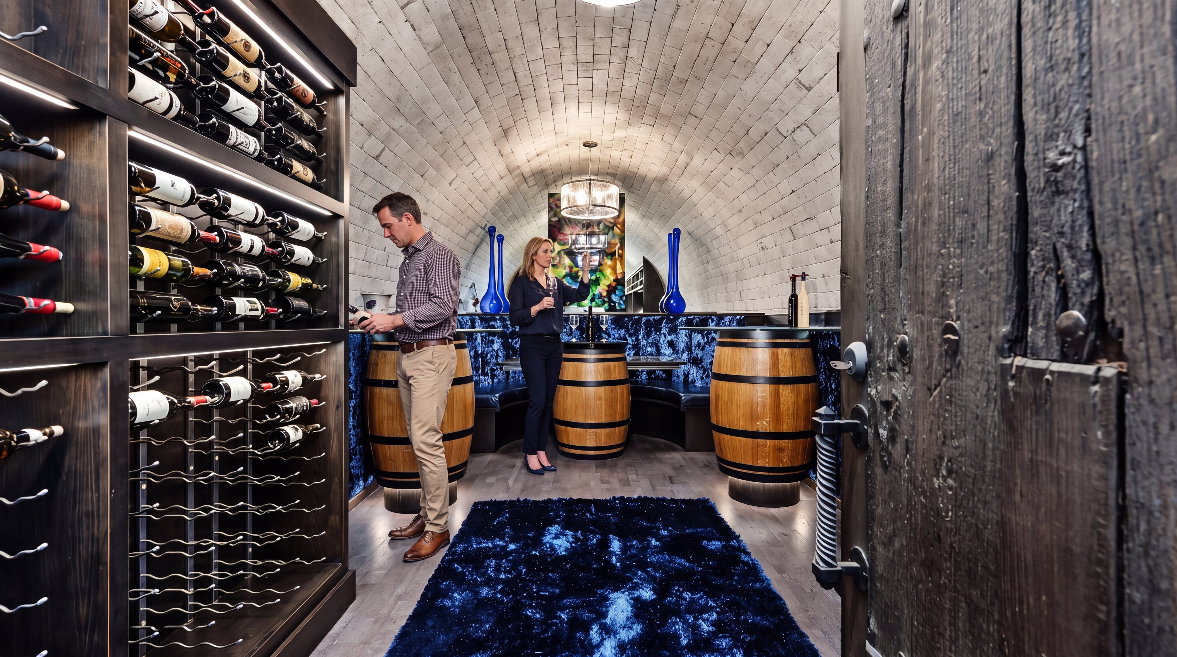 This sophisticated wine cellar features a dramatic barrel-vaulted ceiling, built-in wooden wine racking, and unique barrel-base tasting tables. The space is accented with luxurious deep blue plush carpeting and integrated accent lighting, creating an intimate and upscale atmosphere for wine enthusiasts. The rustic wooden door and stone-textured ceiling add an elegant, character-filled charm to this refined entertaining area.
