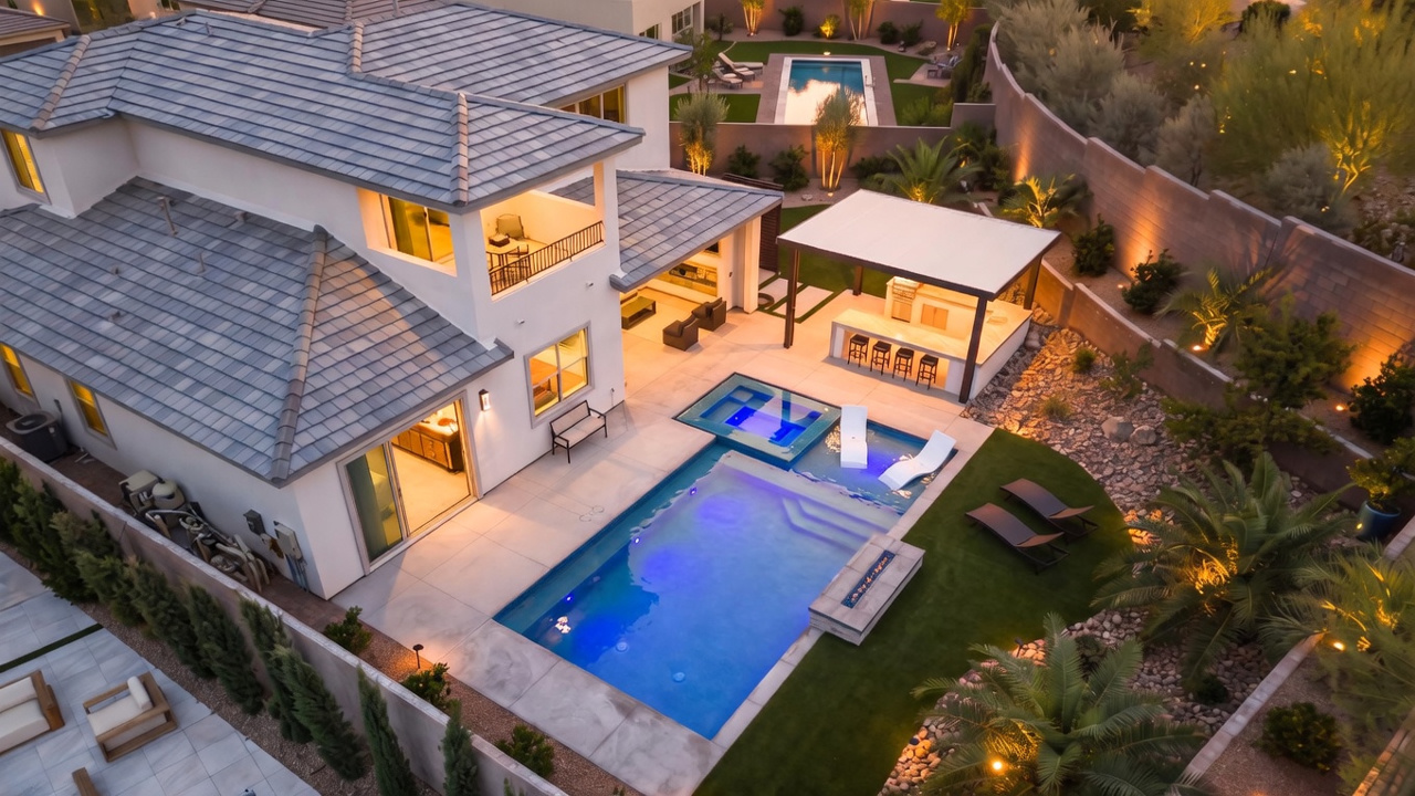 This high-angle aerial view captures a luxurious backyard oasis featuring a modern swimming pool with an integrated spa and sun shelf. A covered outdoor kitchen area with bar seating provides a perfect space for entertaining, while manicured landscaping and ambient evening lighting enhance the sophisticated atmosphere. The property showcases a contemporary architectural style with clean lines and a seamless transition between indoor and outdoor living spaces.