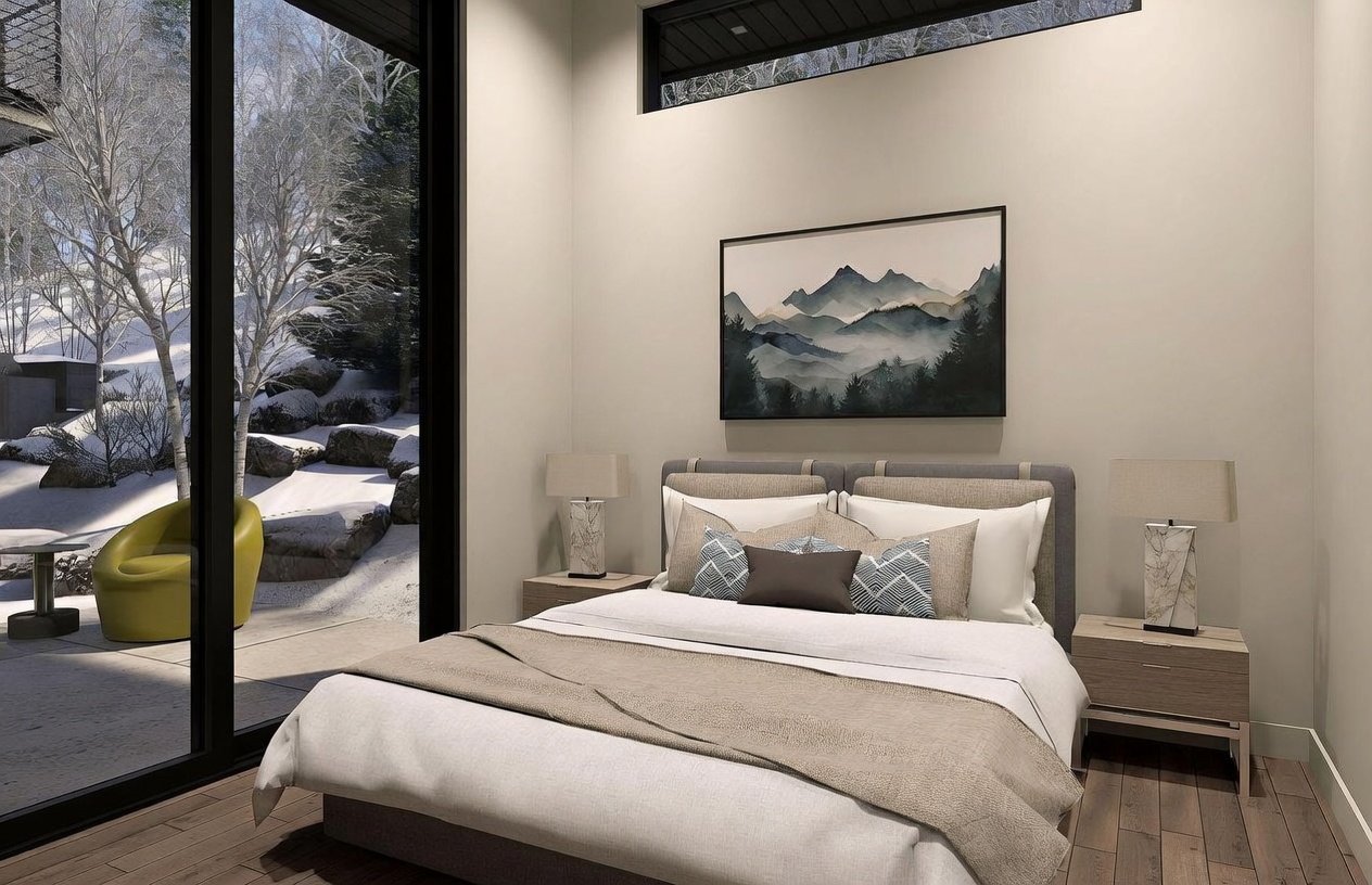 This contemporary bedroom features a neutral color palette with a plush bed dressed in layered linens and decorative pillows. A large glass sliding door provides a seamless view of a snow-covered outdoor landscape, while a piece of mountain-themed artwork hangs above the headboard. The room is accented by two matching bedside lamps on light wood nightstands, creating a serene and modern atmosphere.