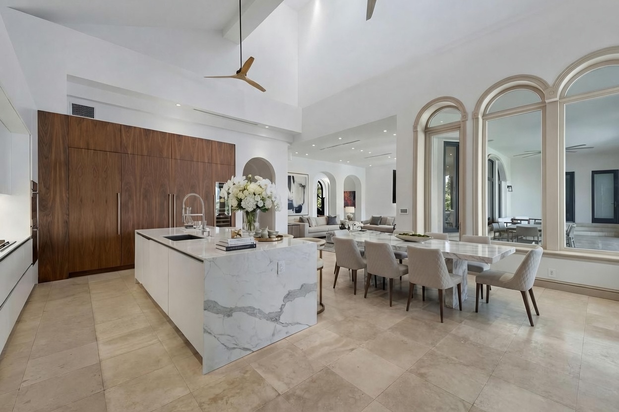 This expansive, open-concept kitchen and dining area features a large central island with a stone waterfall edge and a matching dining table nearby. The space is defined by high vaulted ceilings, dark wood cabinetry, and neutral-toned tile flooring, creating a bright and airy atmosphere. Large arched mirrors reflect the adjacent living space, enhancing the sense of depth and luxury in this modern home.