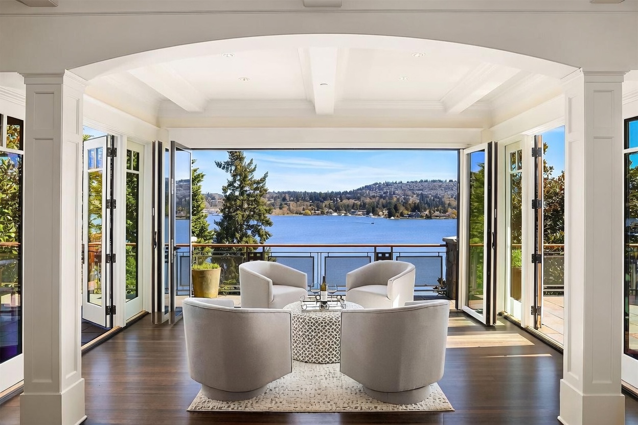 This elegant living space features a sophisticated seating arrangement with four plush, light-gray swivel chairs centered around a patterned circular ottoman. The room is defined by expansive, floor-to-ceiling folding glass doors that open completely to a private balcony, seamlessly blending the interior with a breathtaking panoramic view of the lake and distant hills. The dark hardwood flooring and crisp white trim create a refined, airy atmosphere perfect for luxury waterfront living.