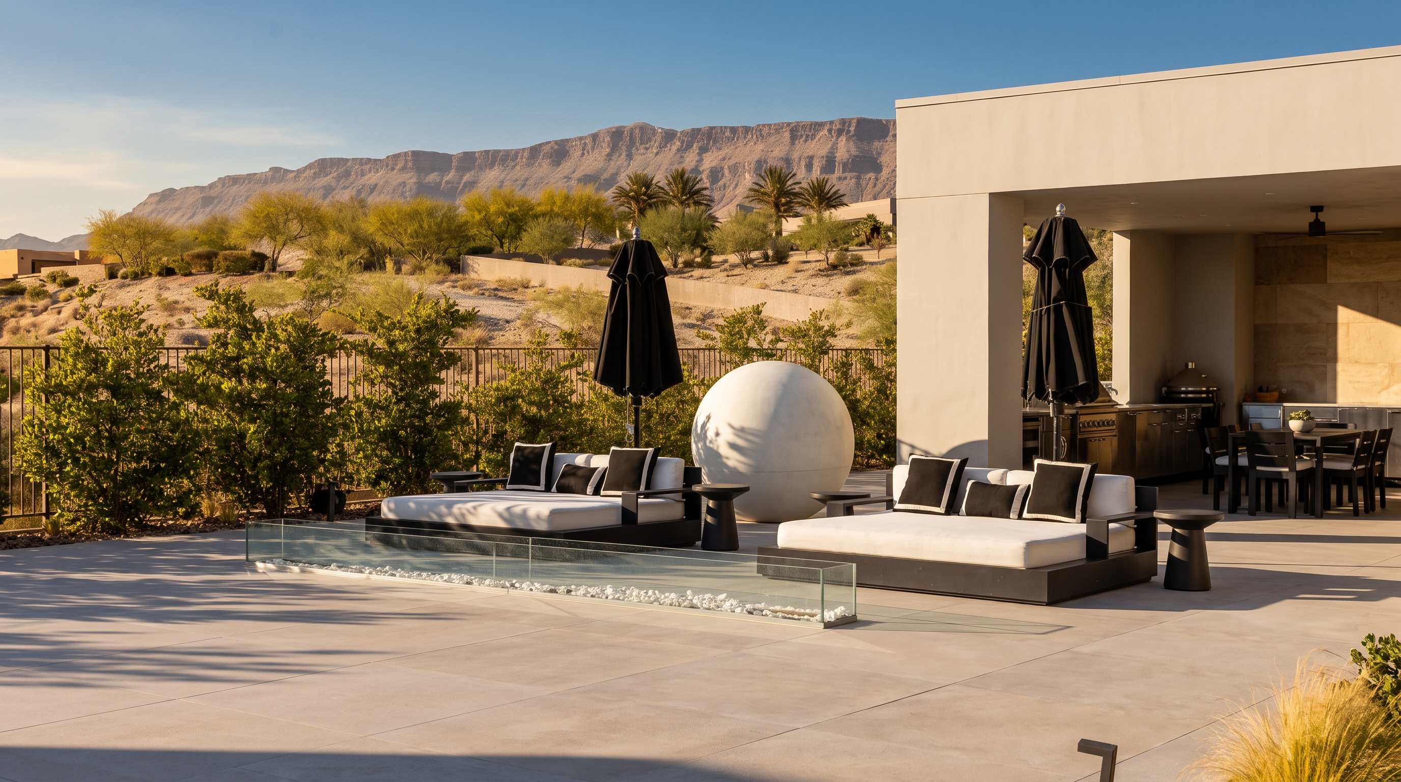 This modern outdoor living space features a sleek infinity-edge pool with a glass-paneled fire feature, set against a backdrop of desert mountains. Two large, contemporary lounge beds with black umbrellas provide ample seating on the expansive concrete patio, which transitions into a covered outdoor kitchen area. The minimalist design emphasizes clean lines and a neutral color palette, creating a sophisticated and serene atmosphere for outdoor entertaining.