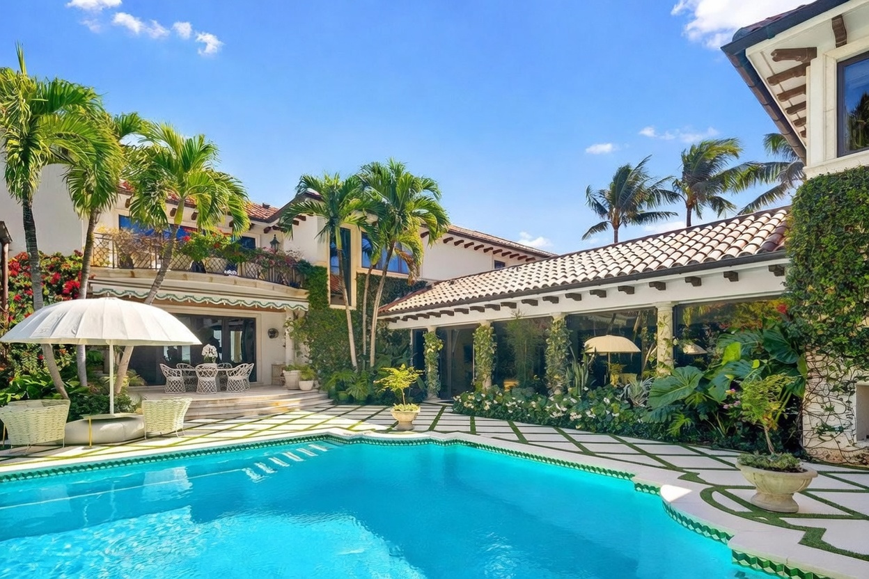 This luxurious Mediterranean-style estate features a stunning backyard oasis centered around a vibrant blue swimming pool. The pool deck is paved with geometric stone tiles, complemented by lush landscaping, tall palm trees, and a covered patio area with outdoor dining furniture. The home's architecture includes warm-toned stucco walls, a terracotta tile roof, and a second-story balcony with decorative iron railings, creating an inviting and sophisticated atmosphere.
