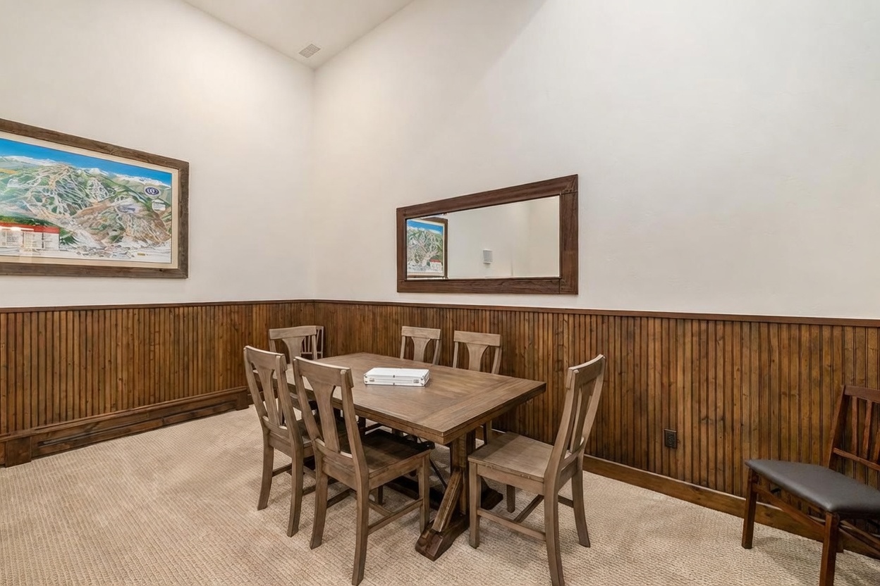 This dining area features a rustic wooden table surrounded by matching chairs, set against a backdrop of warm wood wainscoting. A large framed map of a ski resort hangs on the left wall, while a rectangular mirror reflects the space on the adjacent wall. The room is illuminated by high ceilings and neutral-toned walls, creating a cozy and functional space for gatherings.