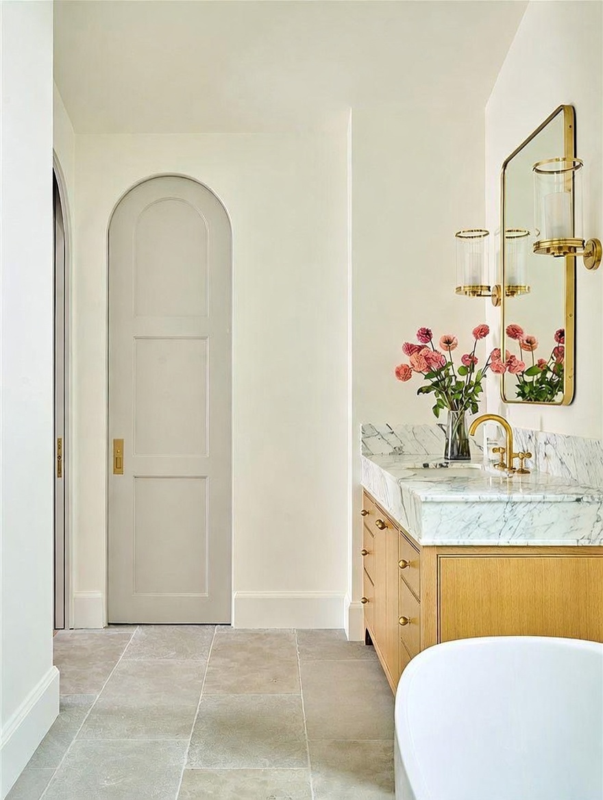 This elegant bathroom features a light wood vanity topped with a stone counter and a gold-toned faucet, complemented by a matching gold-framed mirror and wall sconce. A soft gray arched door adds architectural interest, while the space is finished with large-format stone floor tiles and a vase of fresh flowers. The perspective captures a serene and sophisticated atmosphere, highlighting the clean lines and high-end finishes of the room.
