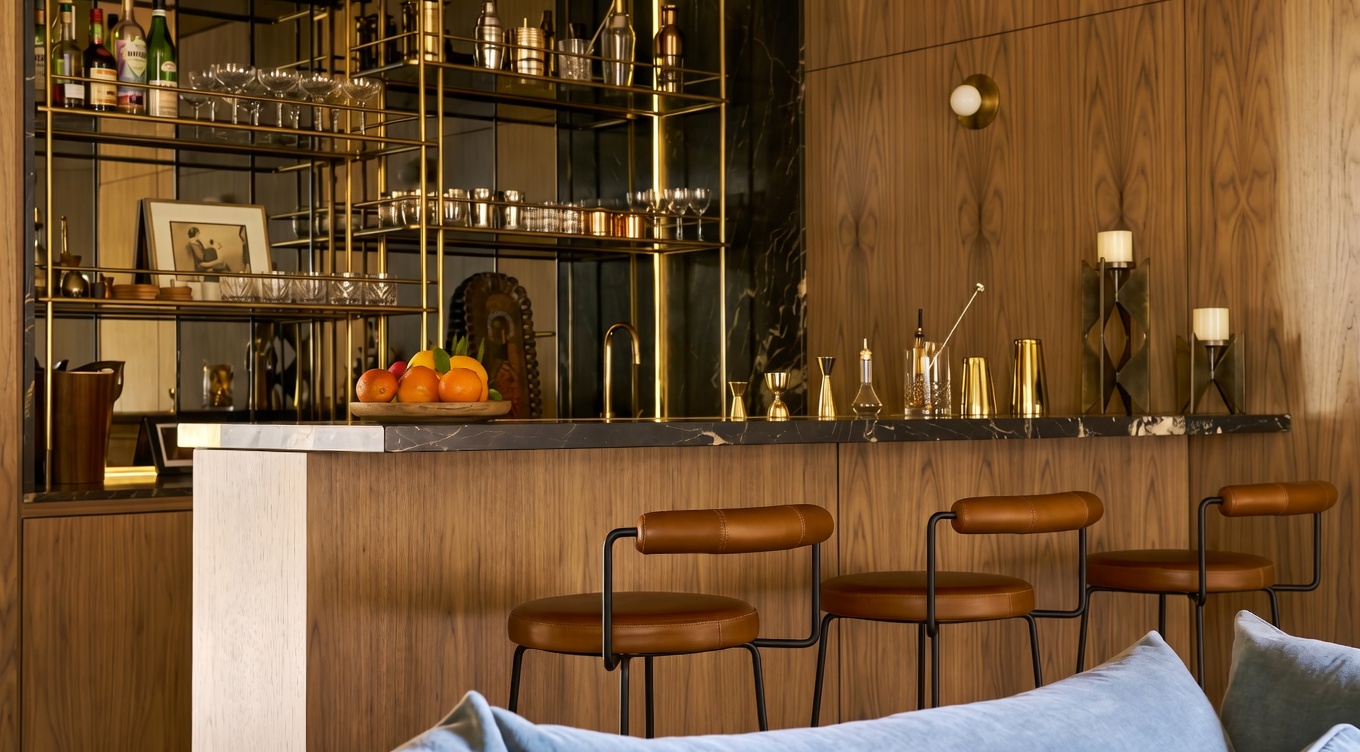 This sophisticated home bar area features warm wood-paneled walls and a sleek stone-topped counter with three leather-upholstered bar stools. Behind the counter, a mirrored shelving unit with integrated lighting displays glassware and bar accessories, creating a refined and moody atmosphere. The foreground shows the back of a plush blue sofa, suggesting an open-concept layout that seamlessly integrates the bar into the living space.