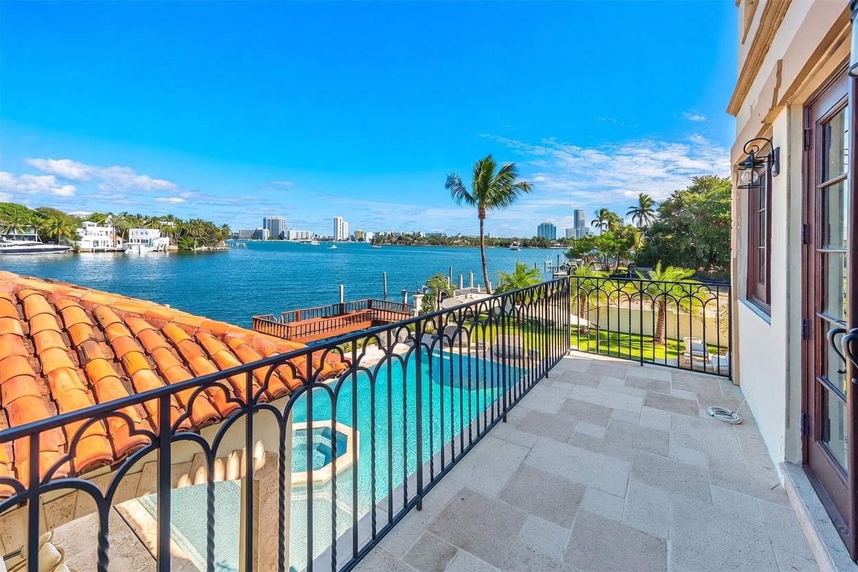 This elevated balcony offers a panoramic view of a tranquil waterway and a distant city skyline under a clear blue sky. The space features light-toned stone flooring, a decorative black metal railing, and a direct view overlooking a private swimming pool and a wooden dock below. The perspective captures the luxurious waterfront lifestyle, highlighting the seamless transition between the home's outdoor living area and the surrounding coastal environment.