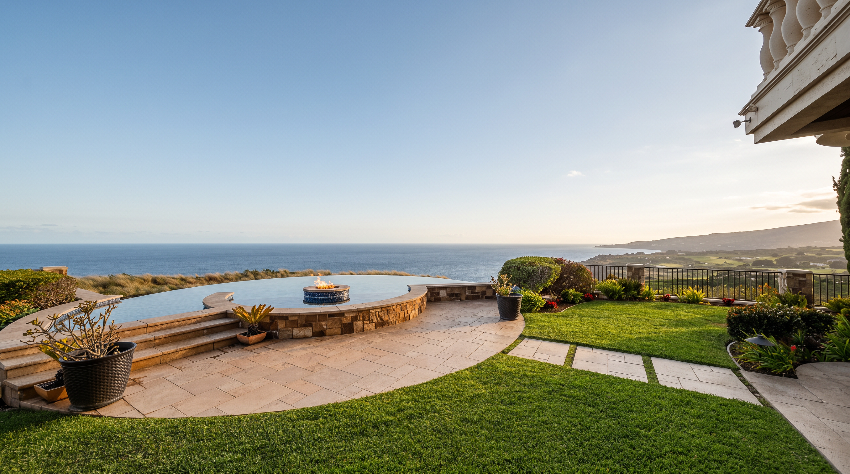 This stunning outdoor space features an infinity-edge pool and integrated spa overlooking a vast, panoramic ocean view. The area includes a paved stone patio, manicured green lawn, and a stone-clad retaining wall, creating a luxurious and serene atmosphere. The perspective captures the seamless transition between the private backyard and the expansive coastal horizon.