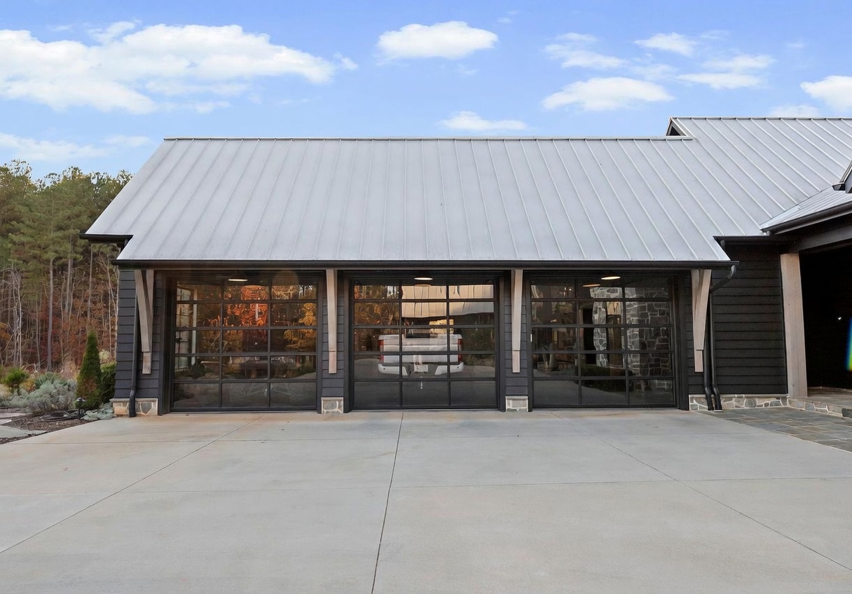 This image showcases a modern three-car garage with a striking industrial aesthetic, featuring dark horizontal siding and a light-colored metal roof. The garage doors are constructed with black frames and large glass panels, providing a contemporary look that reflects the surrounding trees and sky. A spacious concrete driveway leads up to the garage, which is accented by decorative wooden brackets under the eaves.